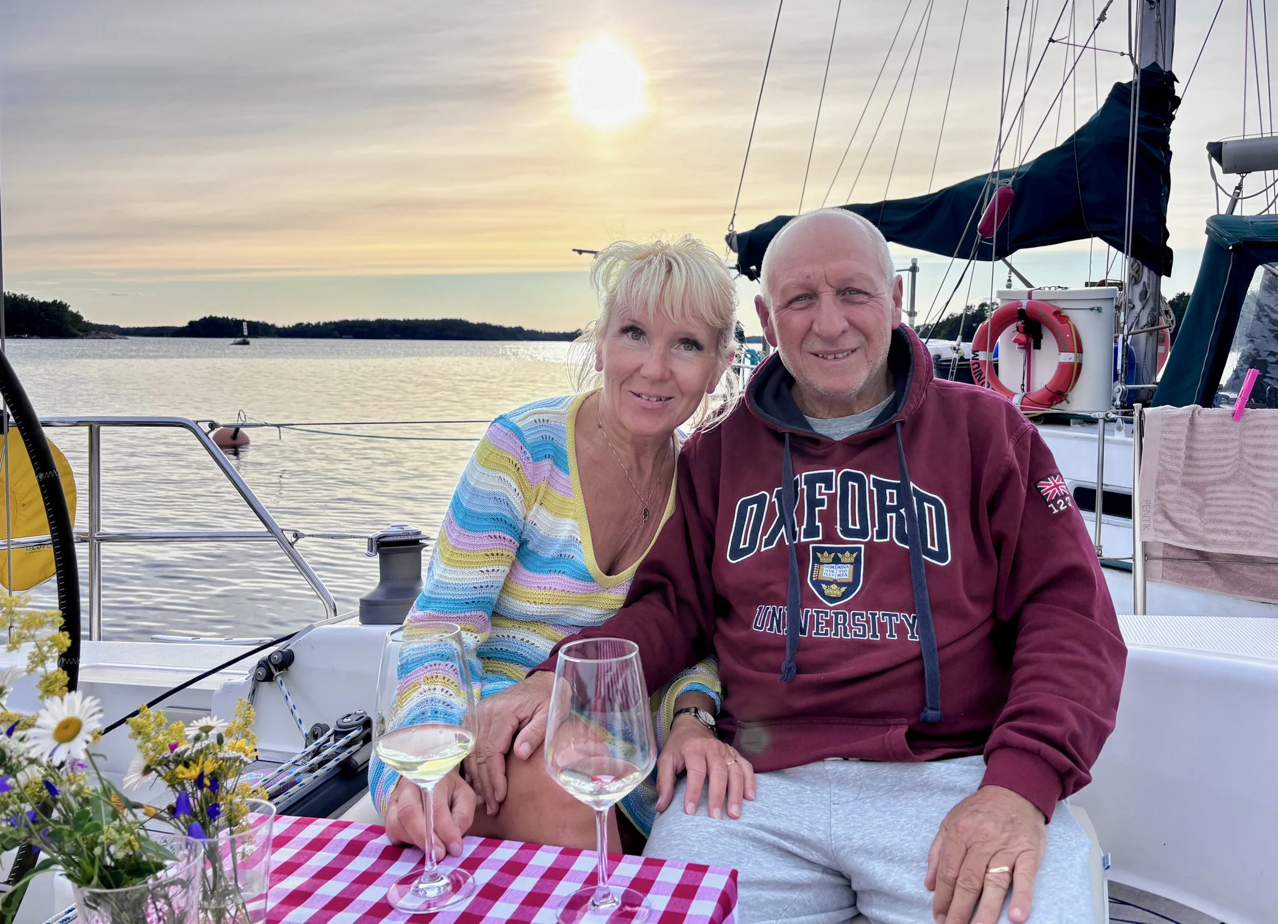 Anita and Umberto sitting at a table with red and white table cloth on a sailing boat, with another boat, sea and sunset in the background