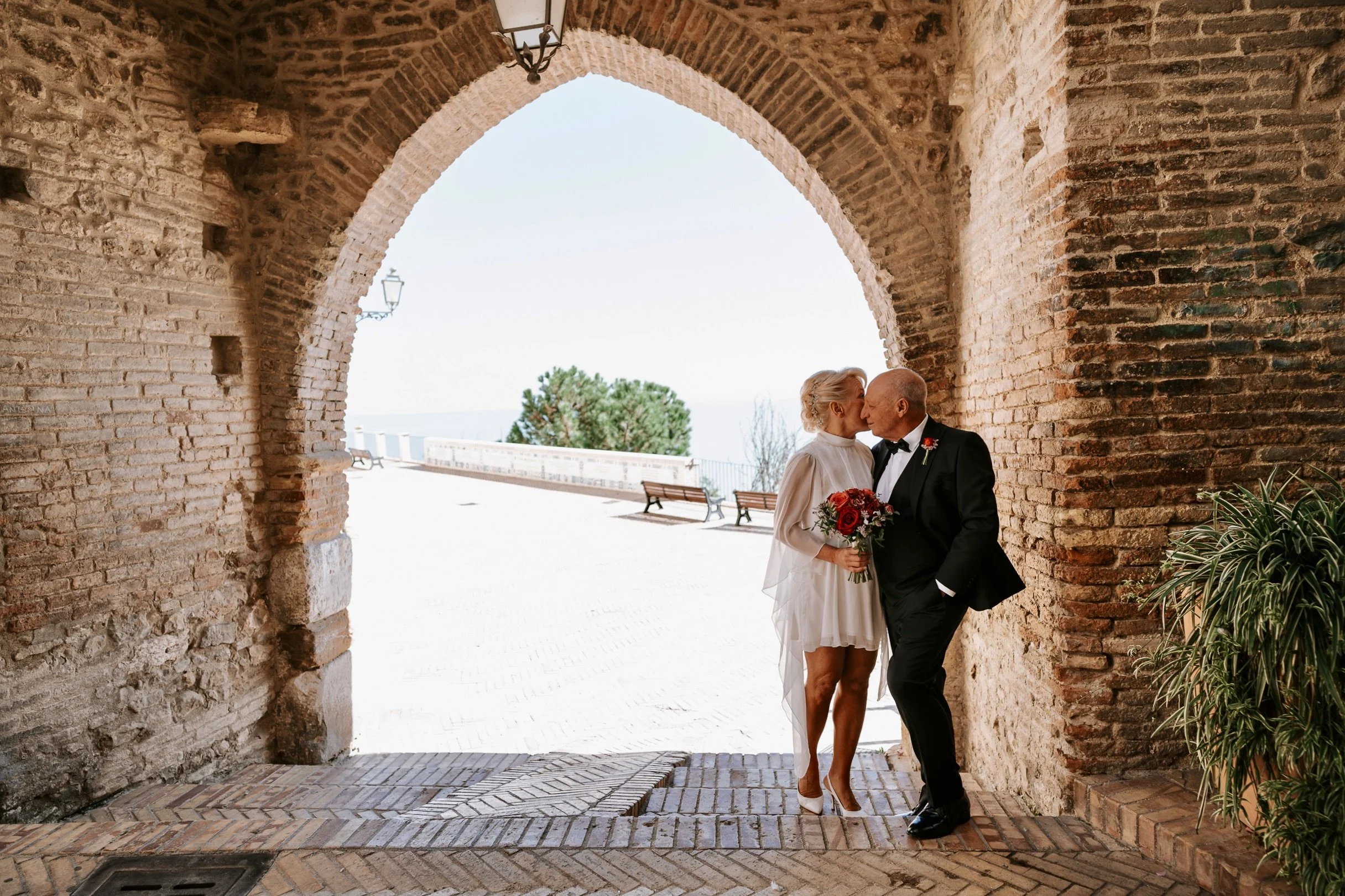 Anita and Umberto in wedding clothes at the Porta Catena gate in Vasto, Italy, with a faint view of the Adriatic sea in the background.