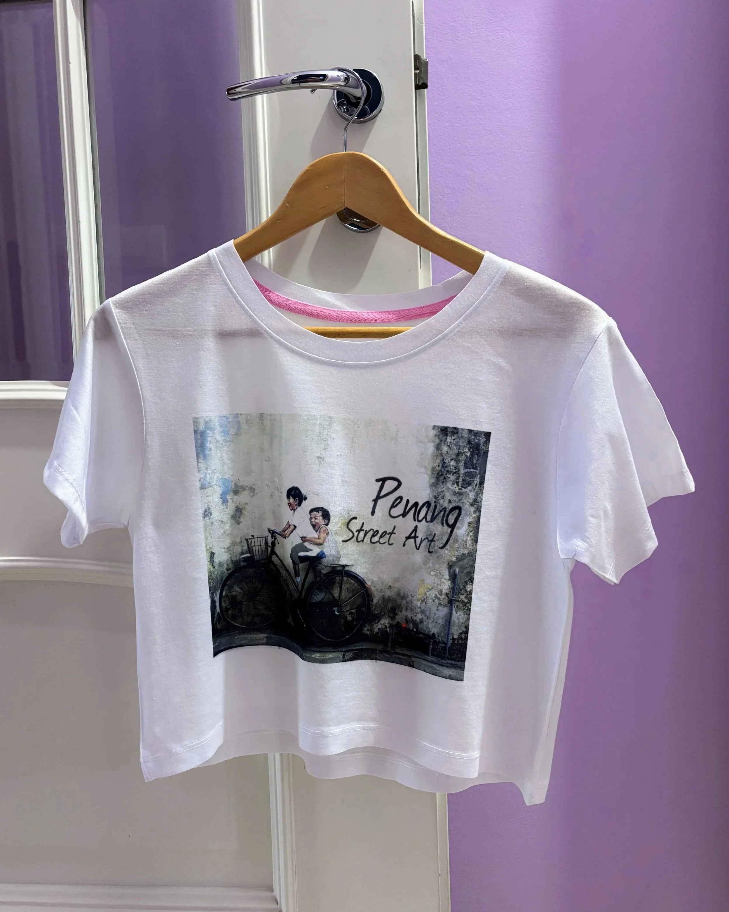 A white t-shirt hanging on a hanger attached to a door handle, the shirt features an illustration of two kids on a bike and the text "Penang Street Art"