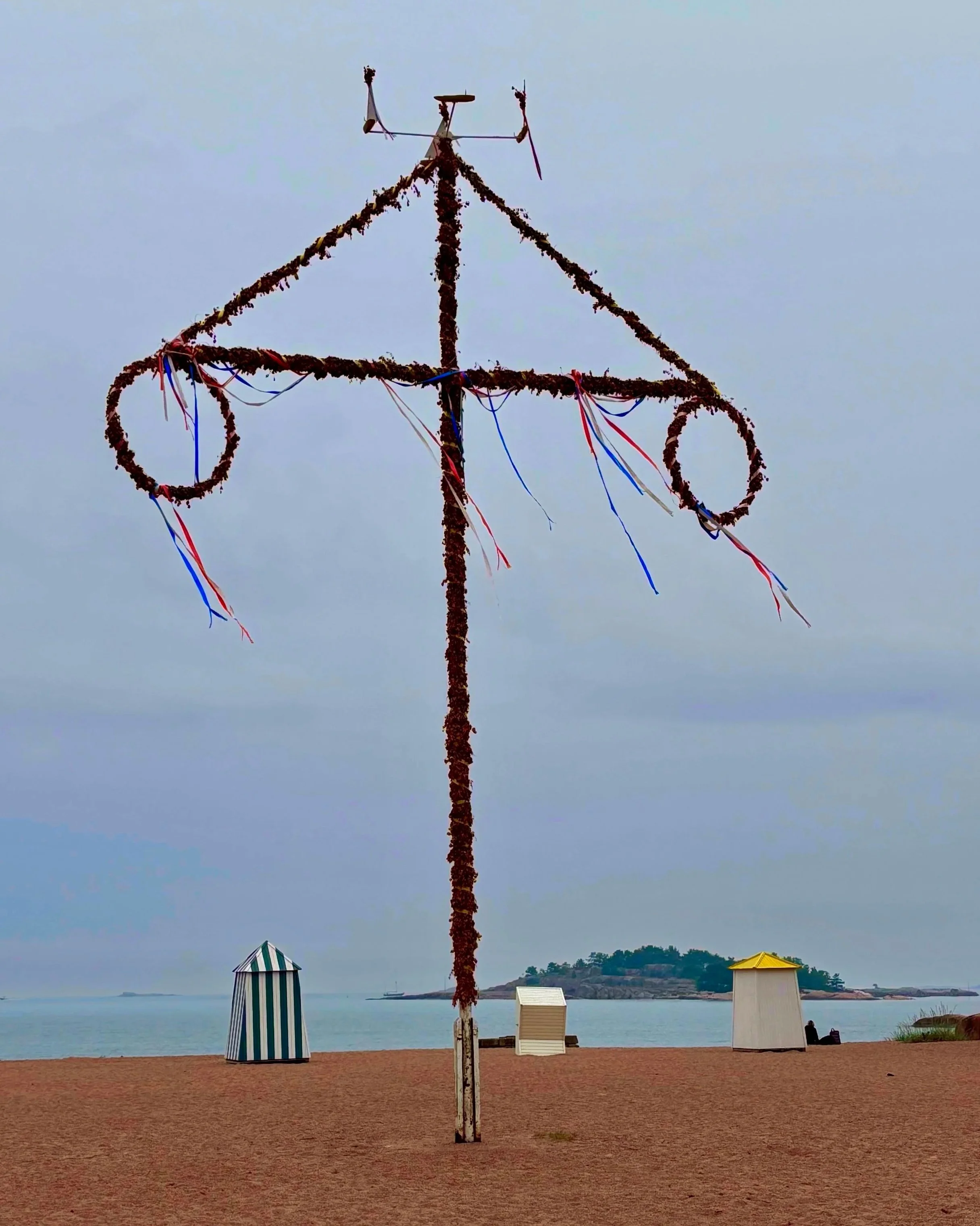 a traditional Midsummer pole on the beach in Hanko, Finland, between two iconic wooden changing cabins
