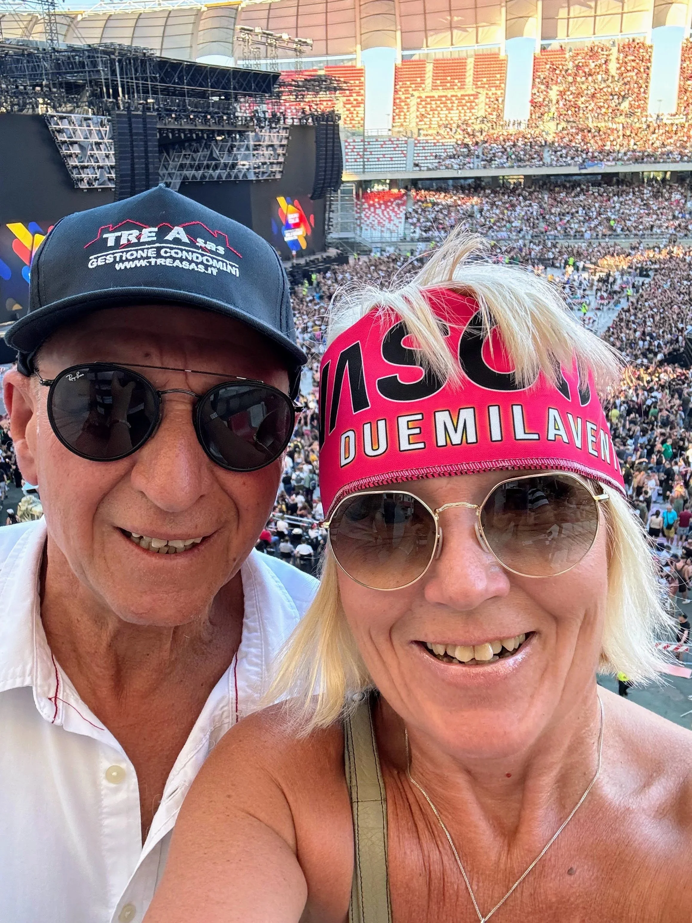 A selfie of Anita and Umberto at the San Nicola stadium in Bari, waiting for the Vasco Rossi concert to begin, with the stage in the background and thousands of fans standing on the field