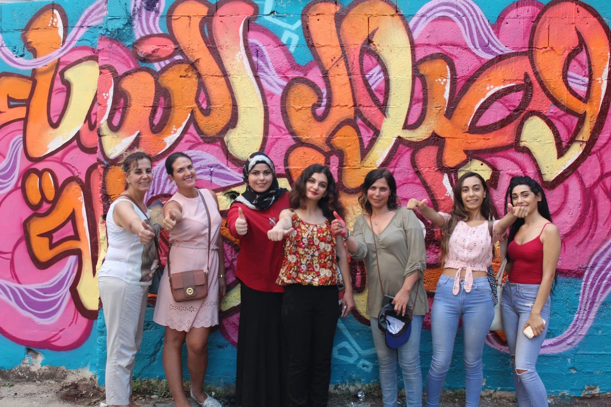 A group of activists standing in front of a graffitied wall hold their hands out with a 'thumbs up' gesture