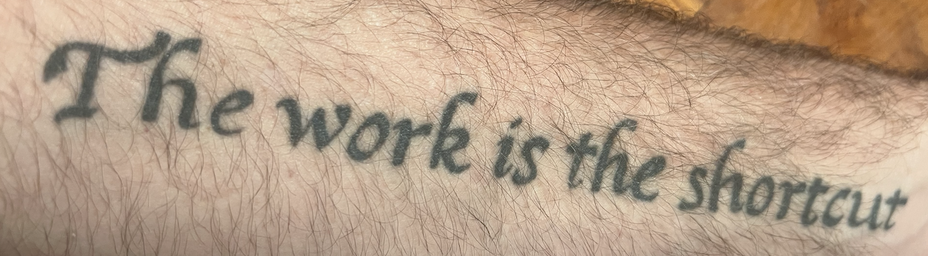 Image of a man's forearm with the words "The work is the shortcut" tattooed on it.