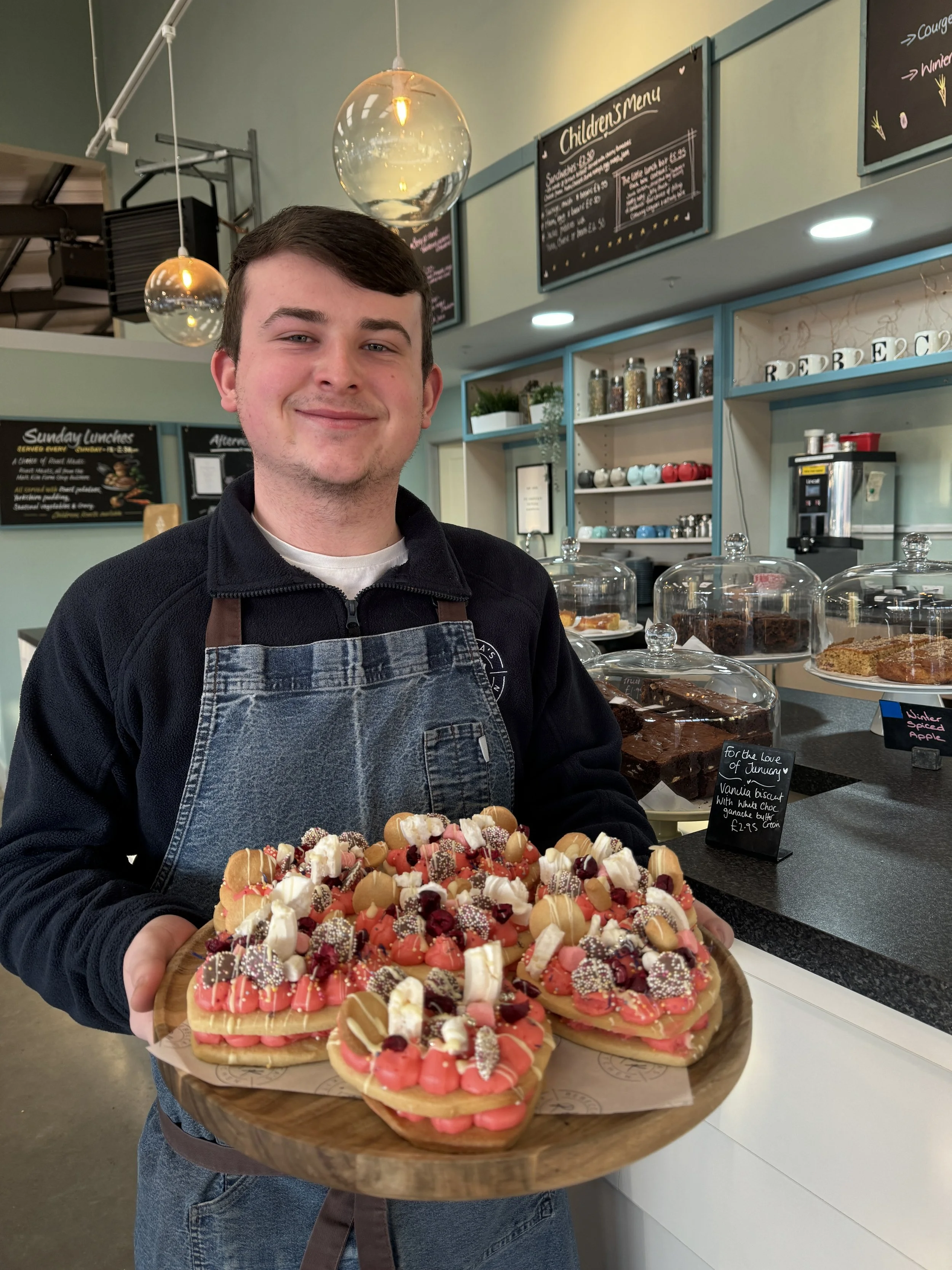Man holding a platter of decorated pastries in a cafe setting, with chalkboard menus and dessert displays in the background.