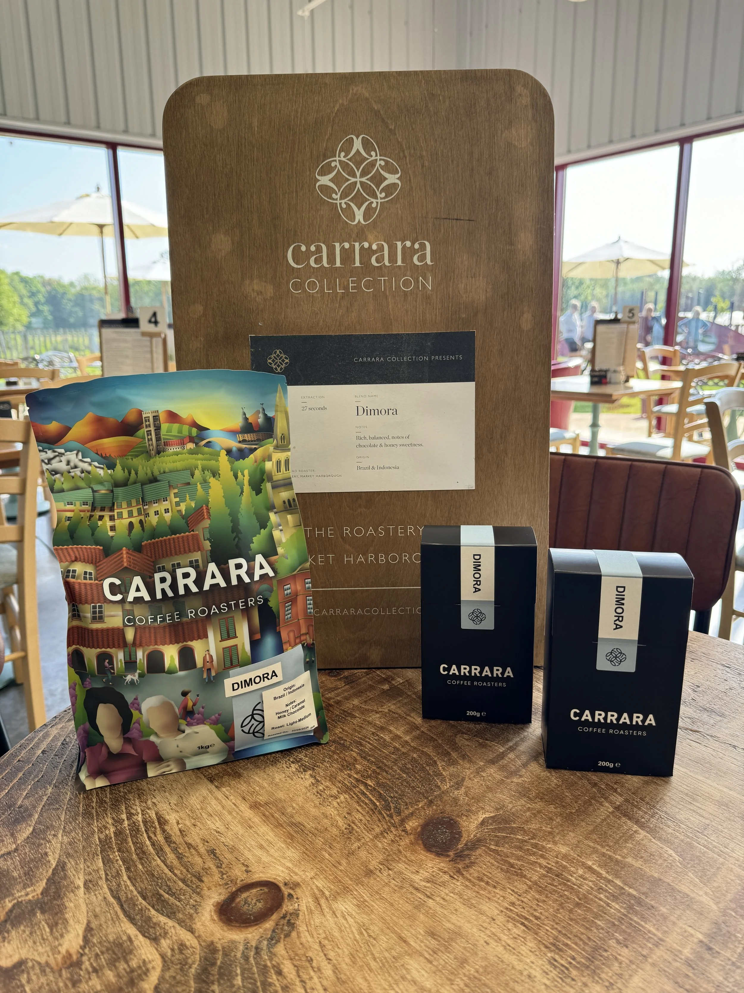 Carrara Collection coffee display on a wooden table, featuring a colorful coffee bag and two black coffee boxes labeled "Dimora," set against a background with a large sign and outdoor seating visible through a window.