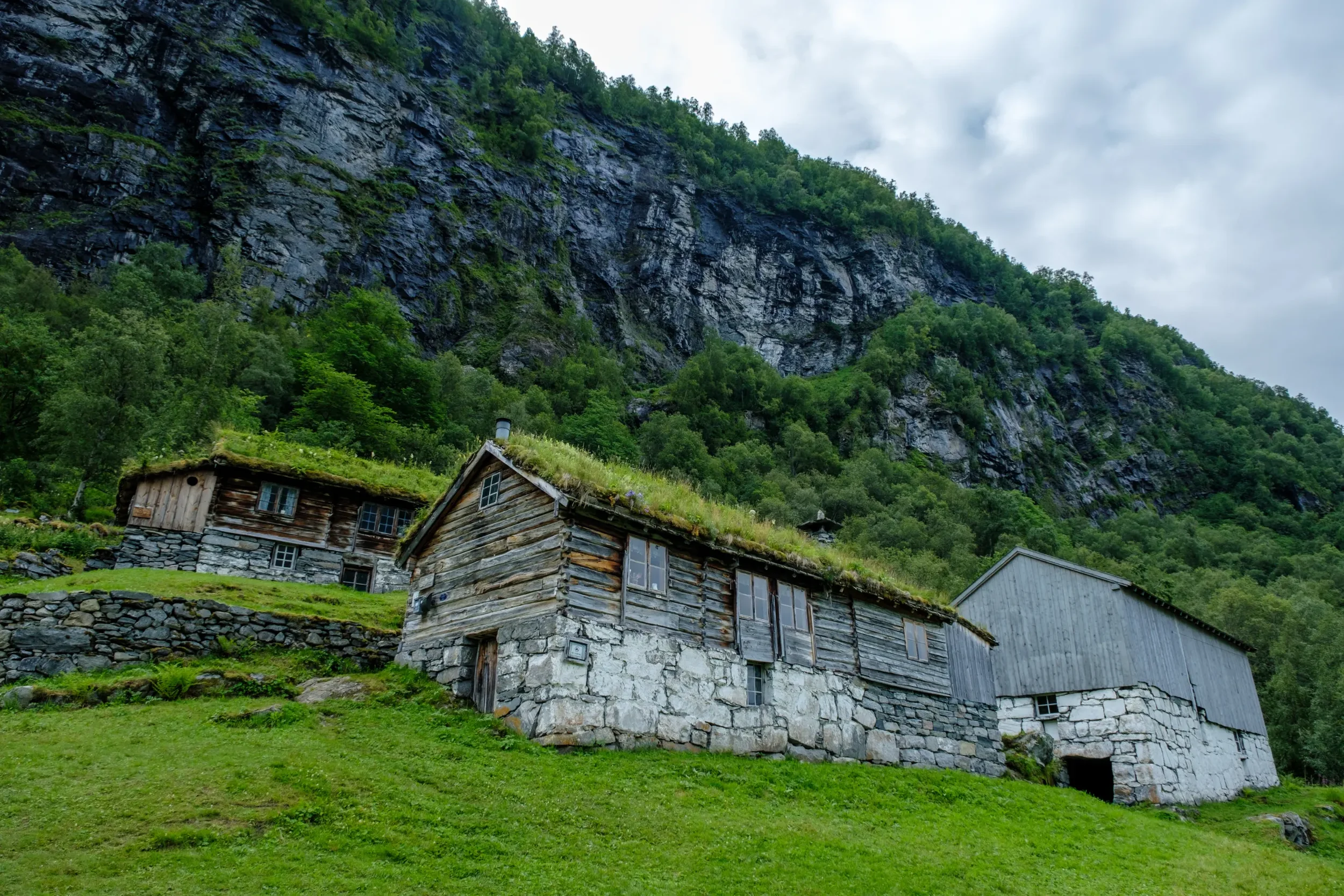 Tre hus med grå stein- og trevegger omgitt av grønt landskap under en fjellskråning