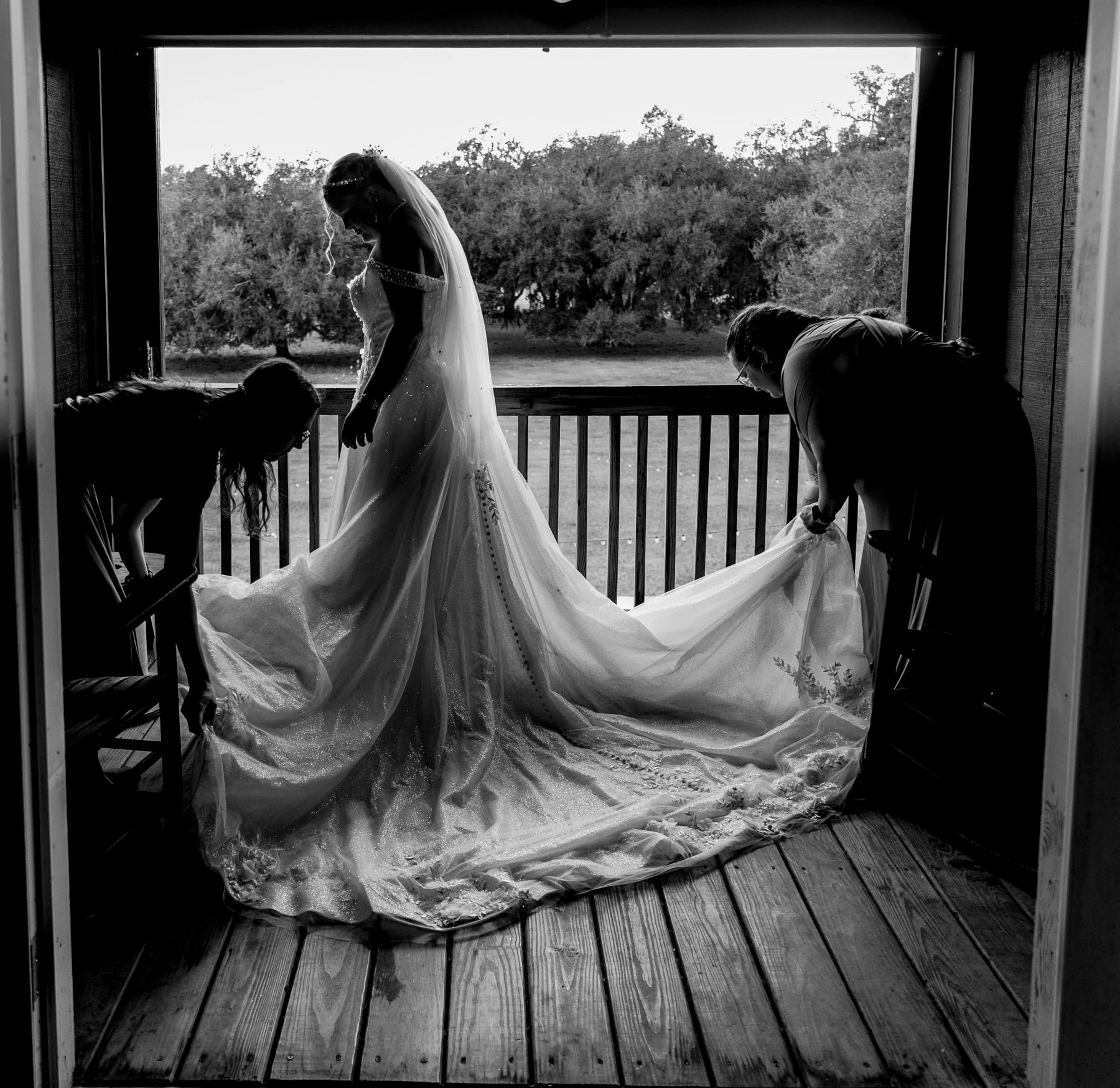 Maddie stands between two of her bridesmaids, backlit on the balcony of the bridal suite. They are adjusting her dress.