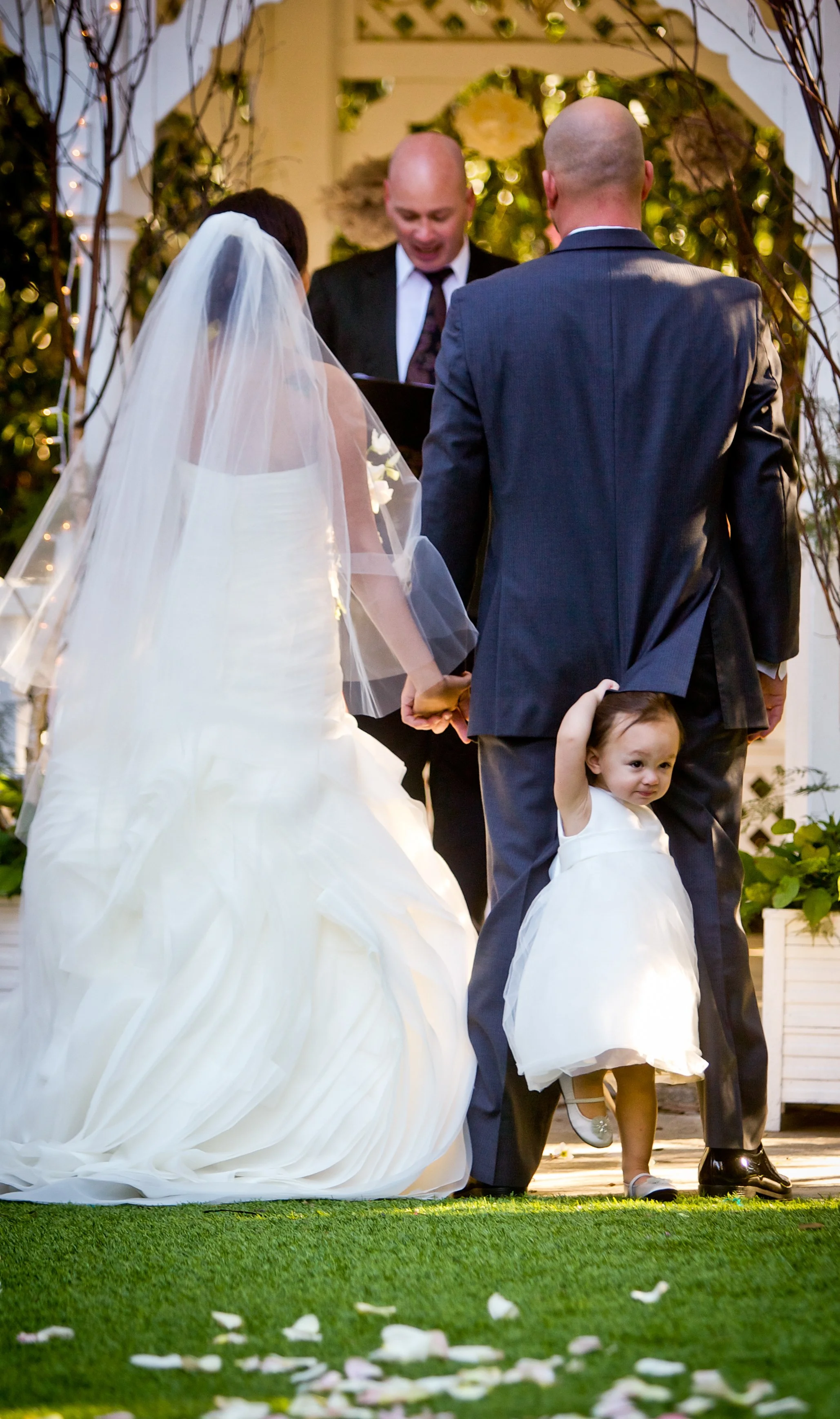 Flower girl holding onto groom’s leg during outdoor wedding ceremony, candid moment showing children at weddings
