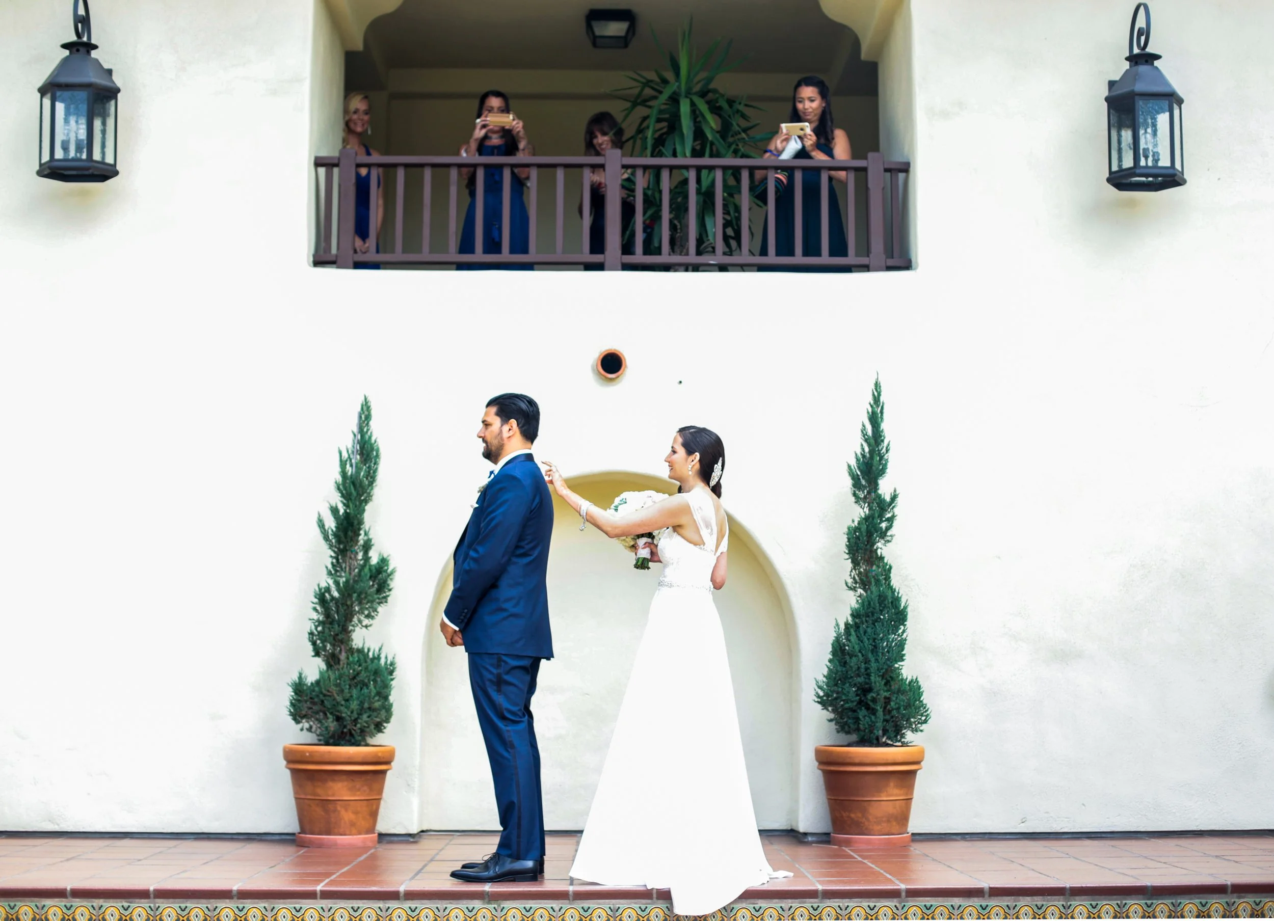 A groom with a blue suit is facing away from his bride as she taps him on the shoulder signifying her can turn around to view her for the first time in her wedding dress. Her bridesmaids are watching above on a balcony and taking photos.