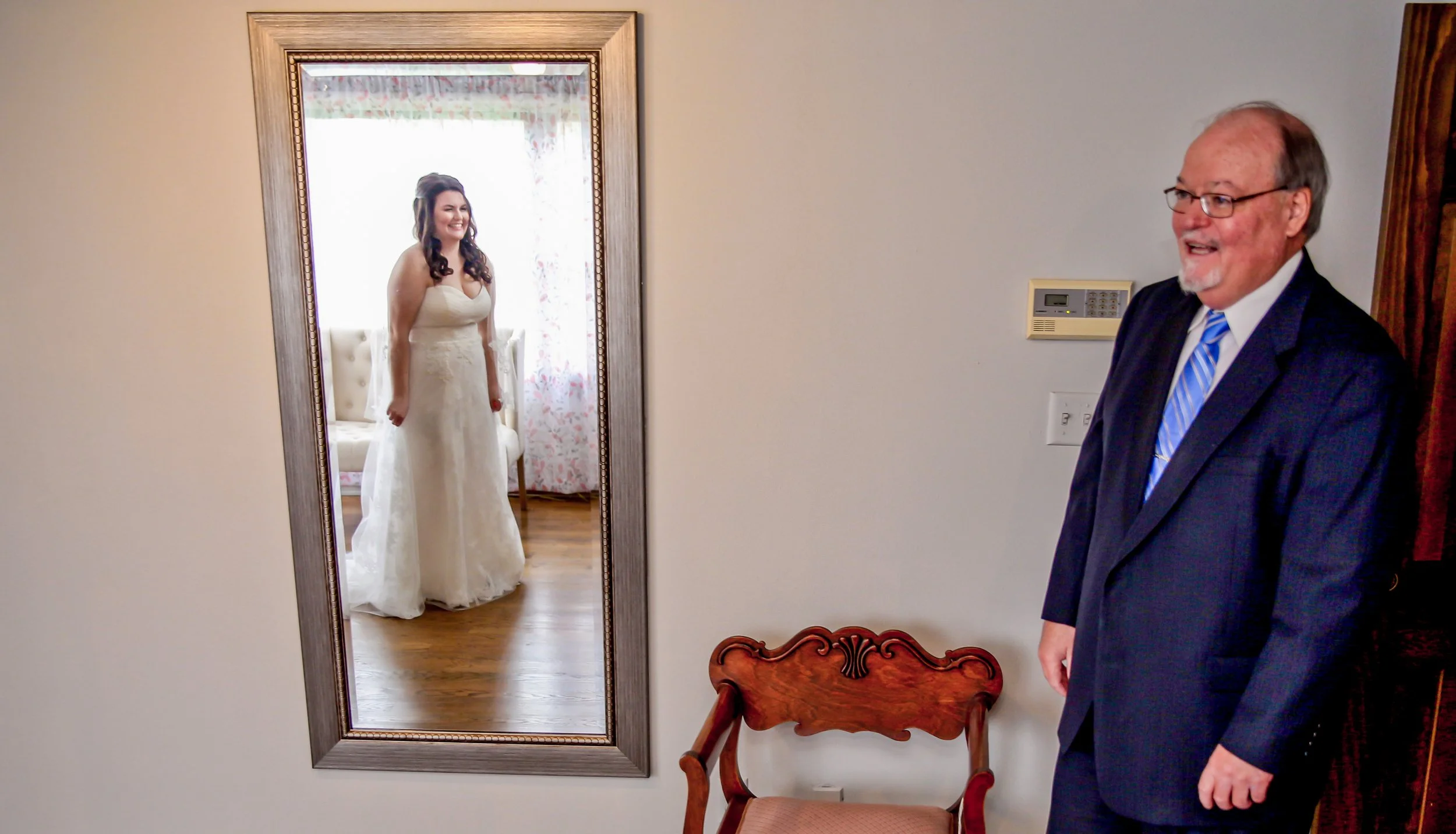 Father of the bride looks at her for the first time after she's gotten ready for her wedding. She's seen in a full-length mirror and he's coming in through the door, smiling.