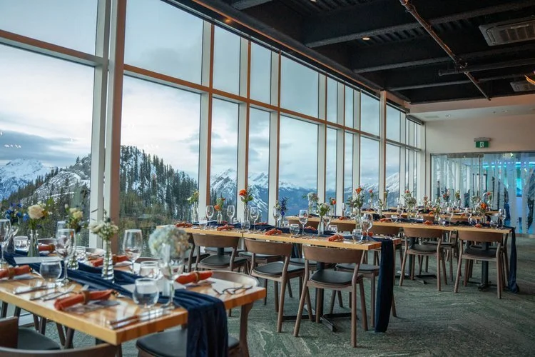Elegant indoor dining area with wooden tables set for a meal, decorated with flowers and glassware, overlooking snow-covered mountains through large floor-to-ceiling windows.