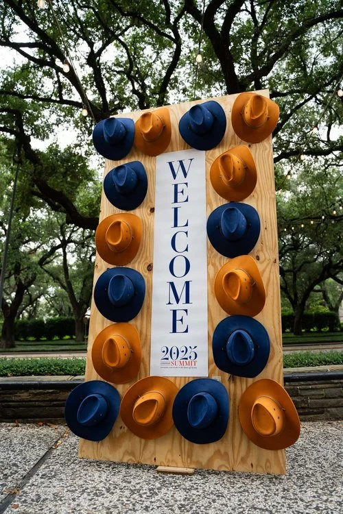 A wooden outdoor welcome sign decorated with blue and orange cowboy hats and a vertical banner that reads "WELCOME 2023 SUMMIT," set against green trees and string lights.