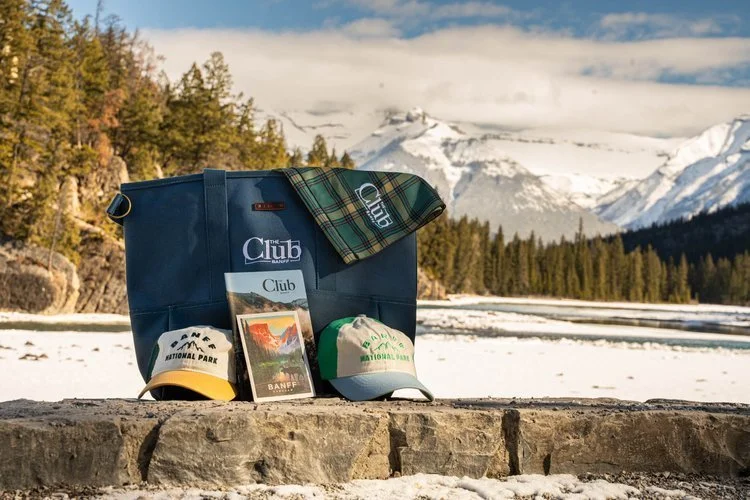 Outdoor scene with a blue bag, a plaid cloth, a yellow and black cap, a green and white cap, and a postcard display in front of a mountain range with snow and a partly cloudy sky.