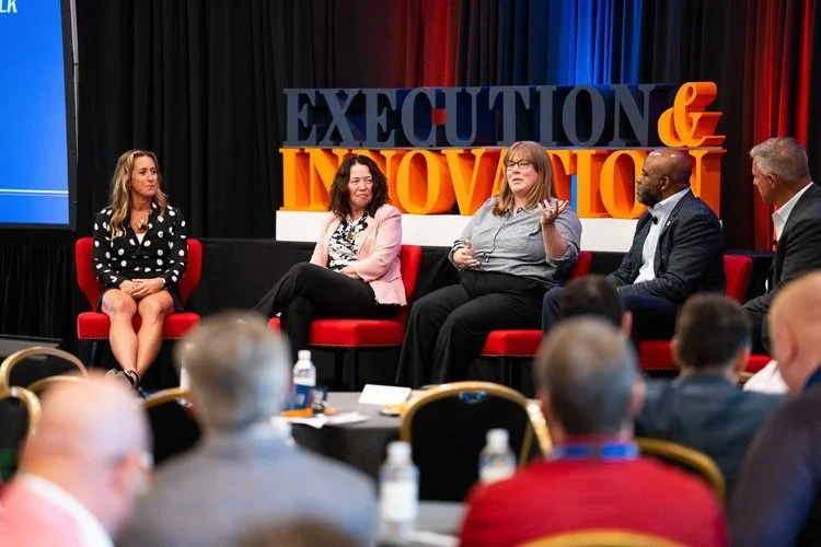 A panel discussion at a conference with five women and one man on stage, seated in red chairs, with a backdrop displaying the words 'Execution & Innovation' in large colorful letters, and an audience listening.
