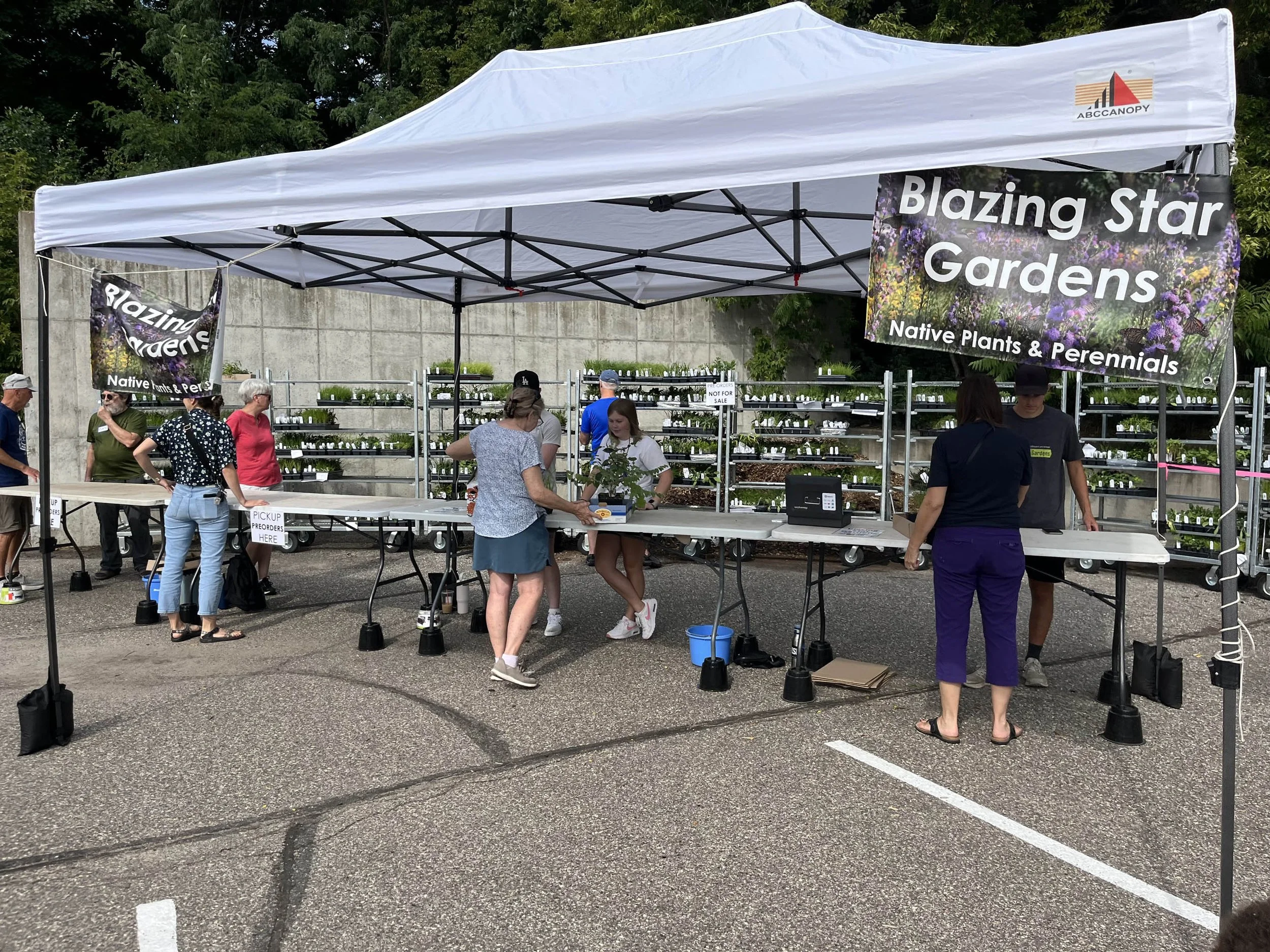 People standing under a canopy in front of metal plant pre-order racks waiting to buy native plants