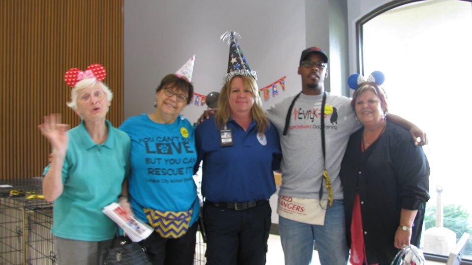 Group of five people wearing playful hats and accessories, standing indoors, smiling, with party decorations in the background.