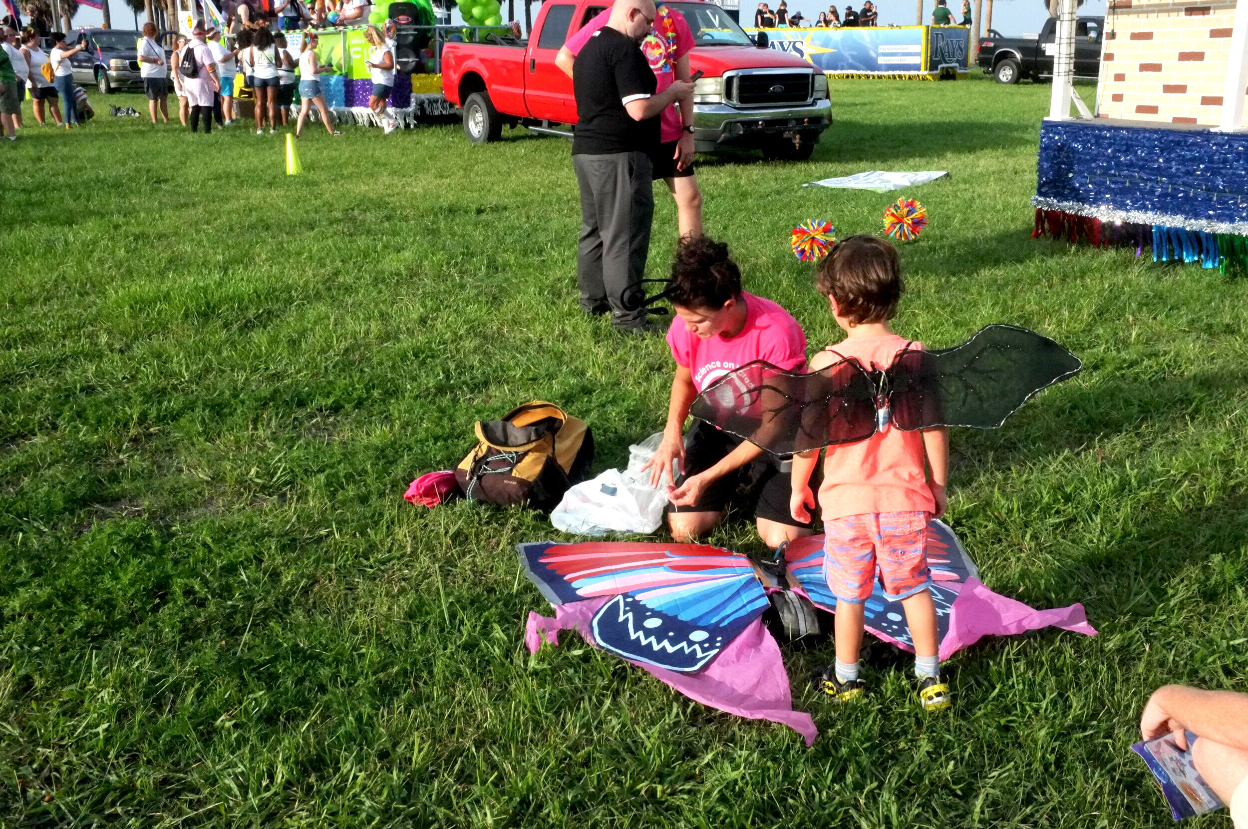  An adult kneeling on the grass preparing wings in the colors of the transgender flag, while a child stands nearby 