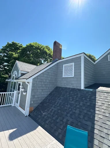 A house with gray siding and a chimney, featuring a deck with white railing and a blue chair, under a clear blue sky with sunlight.