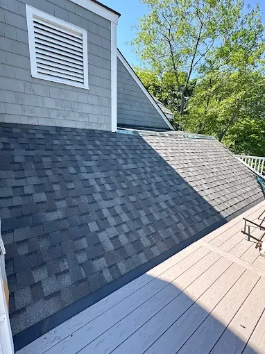 The image shows a rooftop with asphalt shingles, part of a house's exterior wall with a vent, and a wooden deck with outdoor furniture, surrounded by green trees under a clear blue sky.