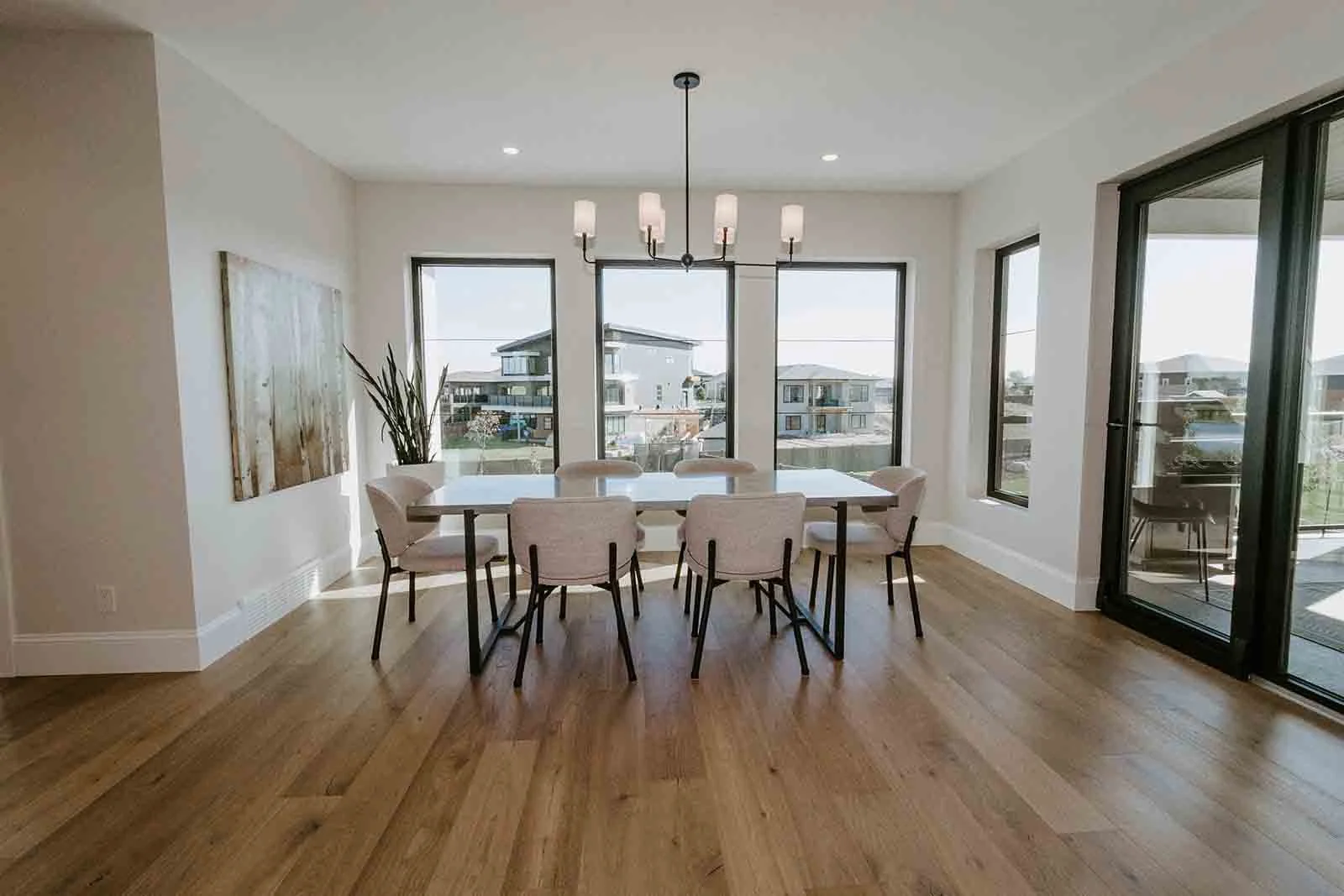 Bright dining room with wooden floor, white walls, a rectangular table with beige chairs, large windows, a chandelier, and a sliding glass door leading to an outdoor balcony.