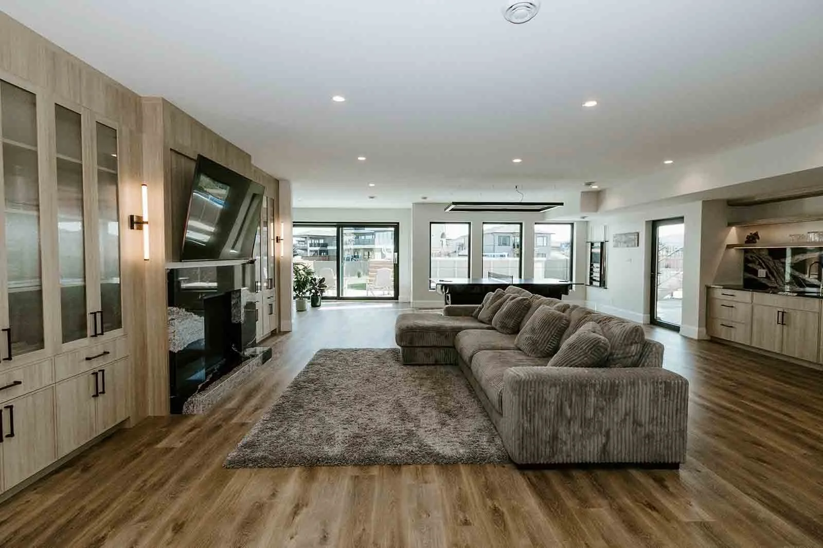 Living room with a large sectional sofa, a plush area rug, built-in wall cabinets, a wall-mounted TV above a fireplace, large windows, a sliding door, and hardwood flooring.