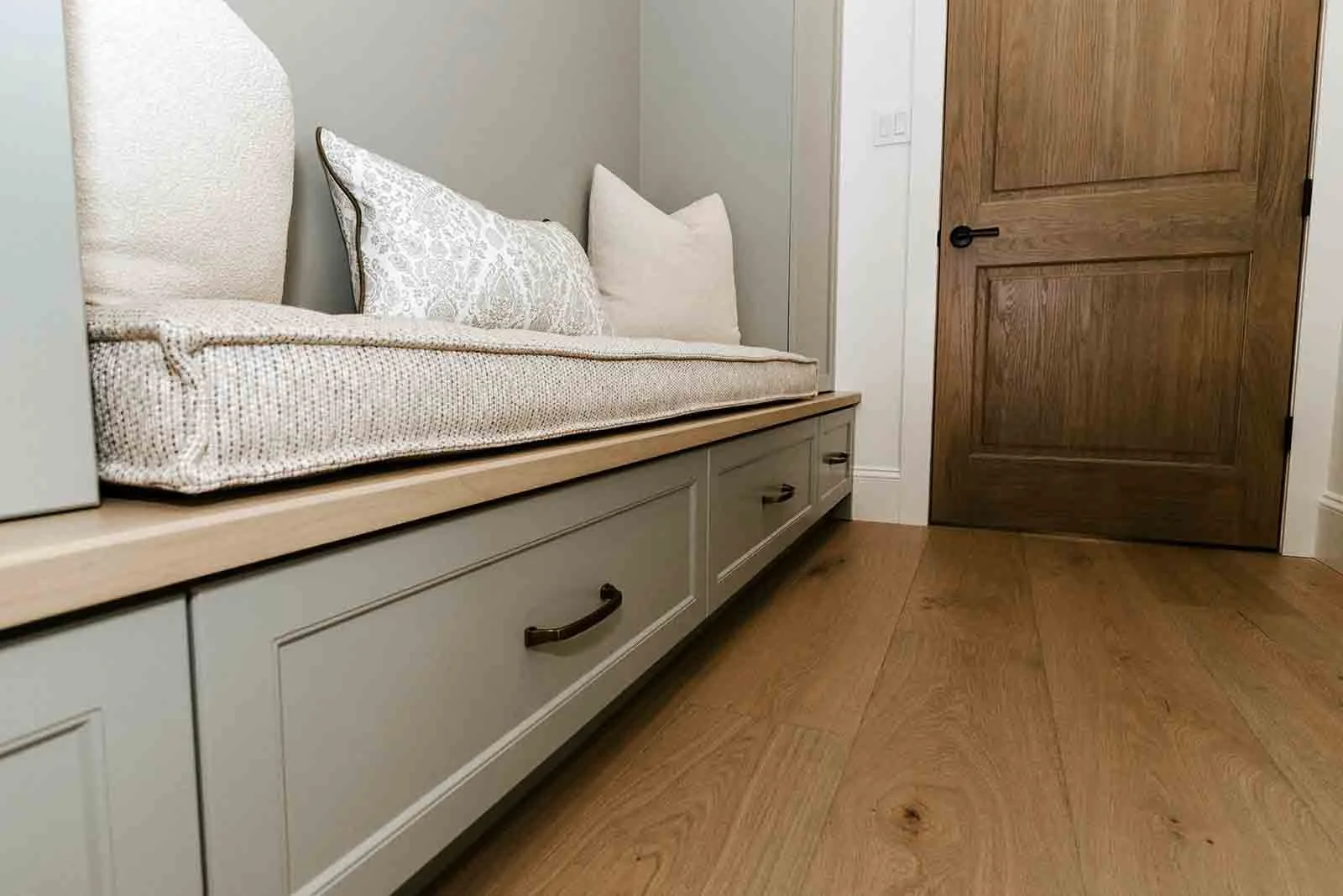 Close-up of a built-in bench with storage drawers beneath, decorated with pillows, next to a wooden door and on a wooden floor.