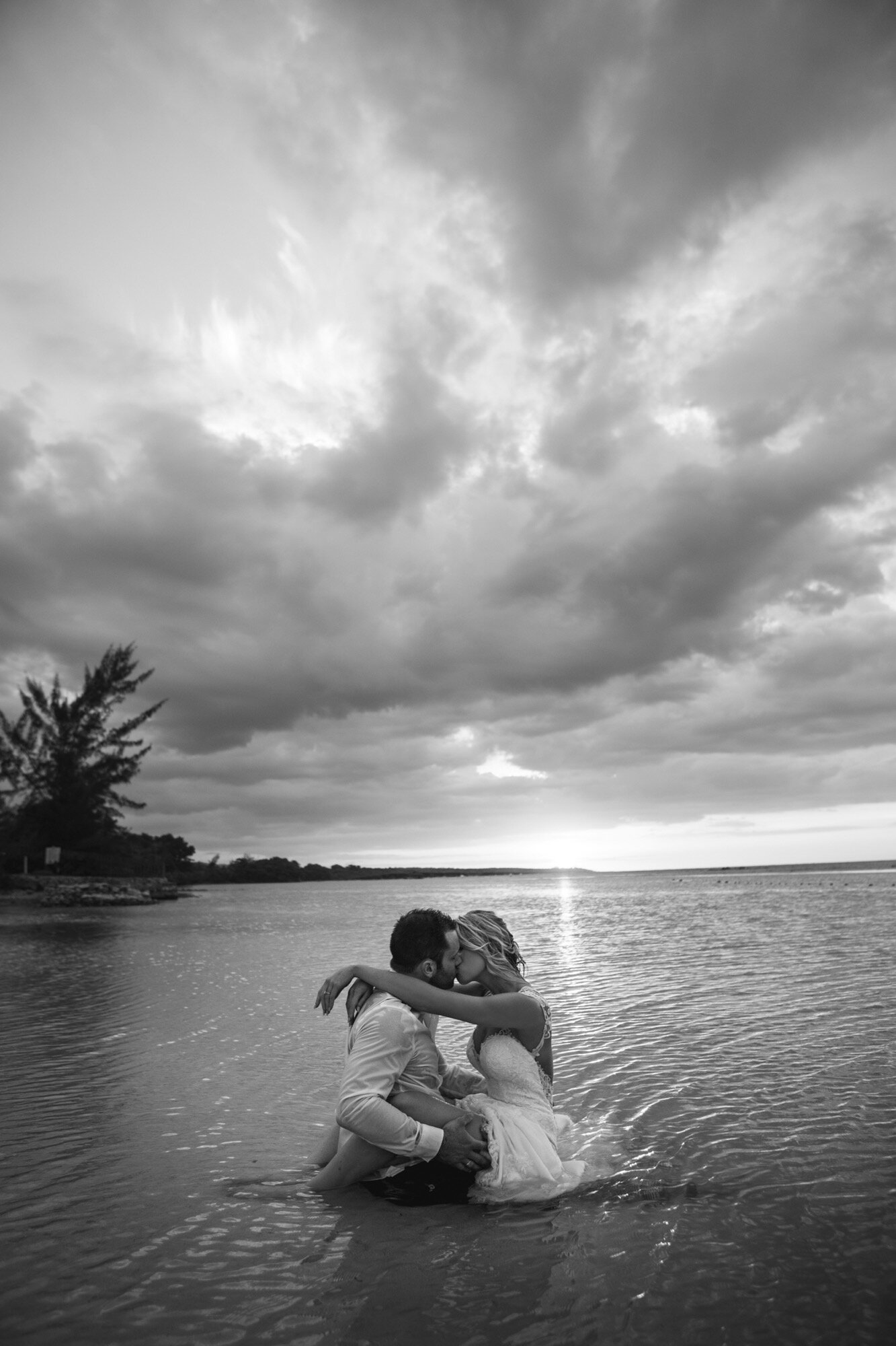 bride groom sitting in ocean kissing at sunset black and white.jpg