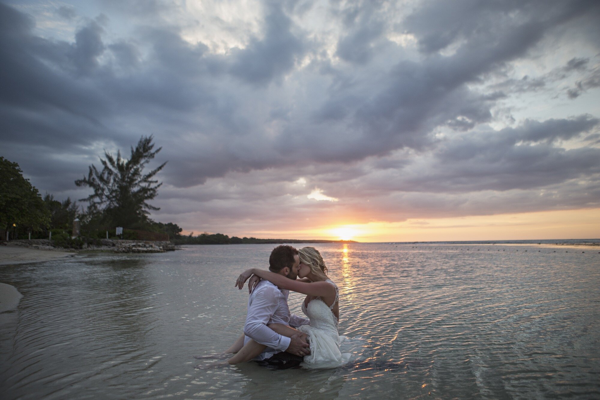 bride groom sitting in shallow water at sunset kissing.jpg