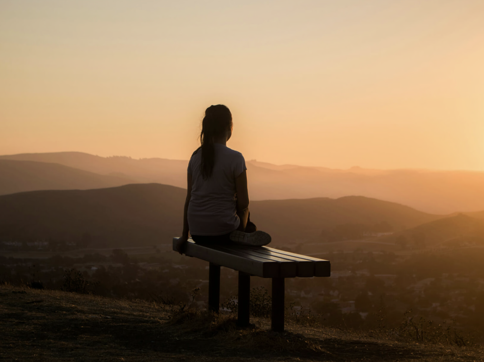 A person sitting on a bench overlooking a landscape with rolling hills during sunset.