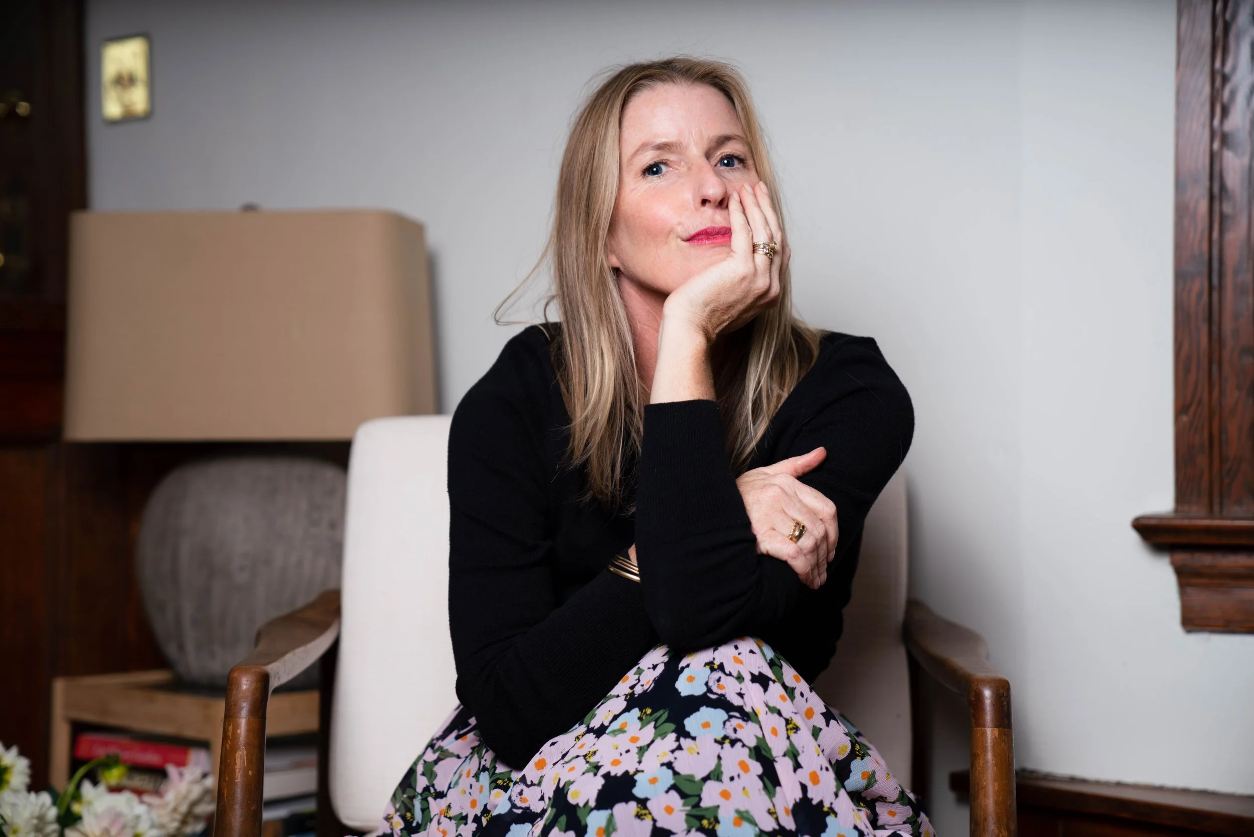 A woman with blonde hair and fair skin sitting on a wooden chair, resting her chin on her hand, looking thoughtfully at the camera inside a room with light-colored walls and furniture.