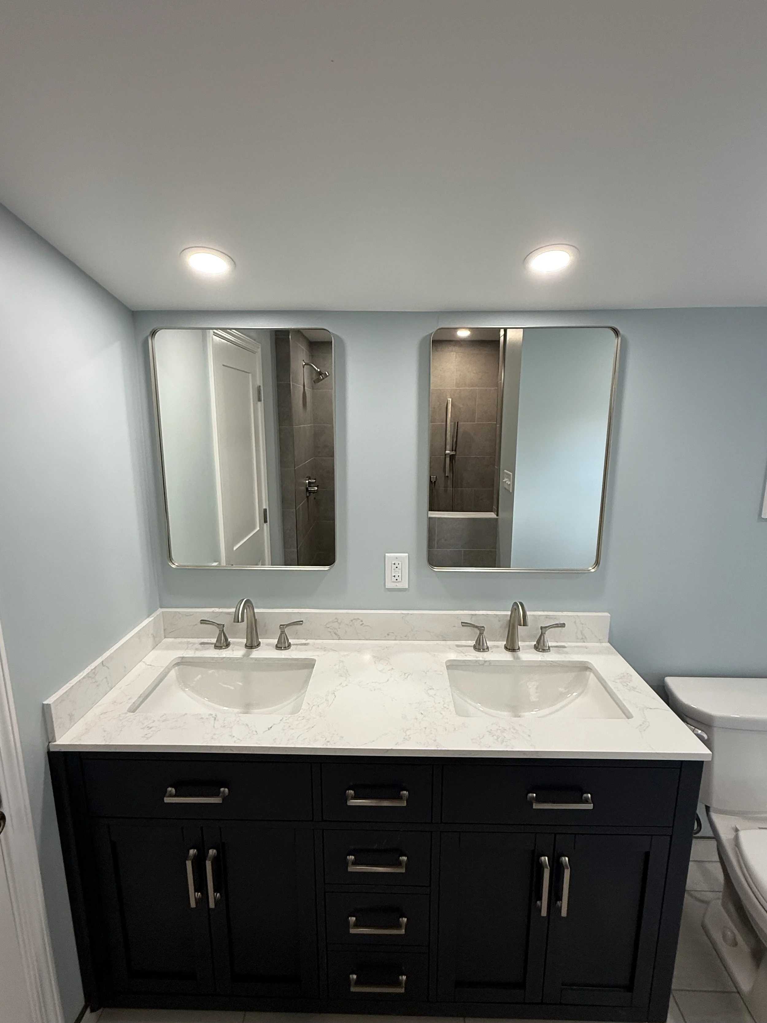 Double vanity bathroom with white marble countertop, two sinks, and two mirrors, with a shower visible in the background, light blue walls, and ceiling lights.