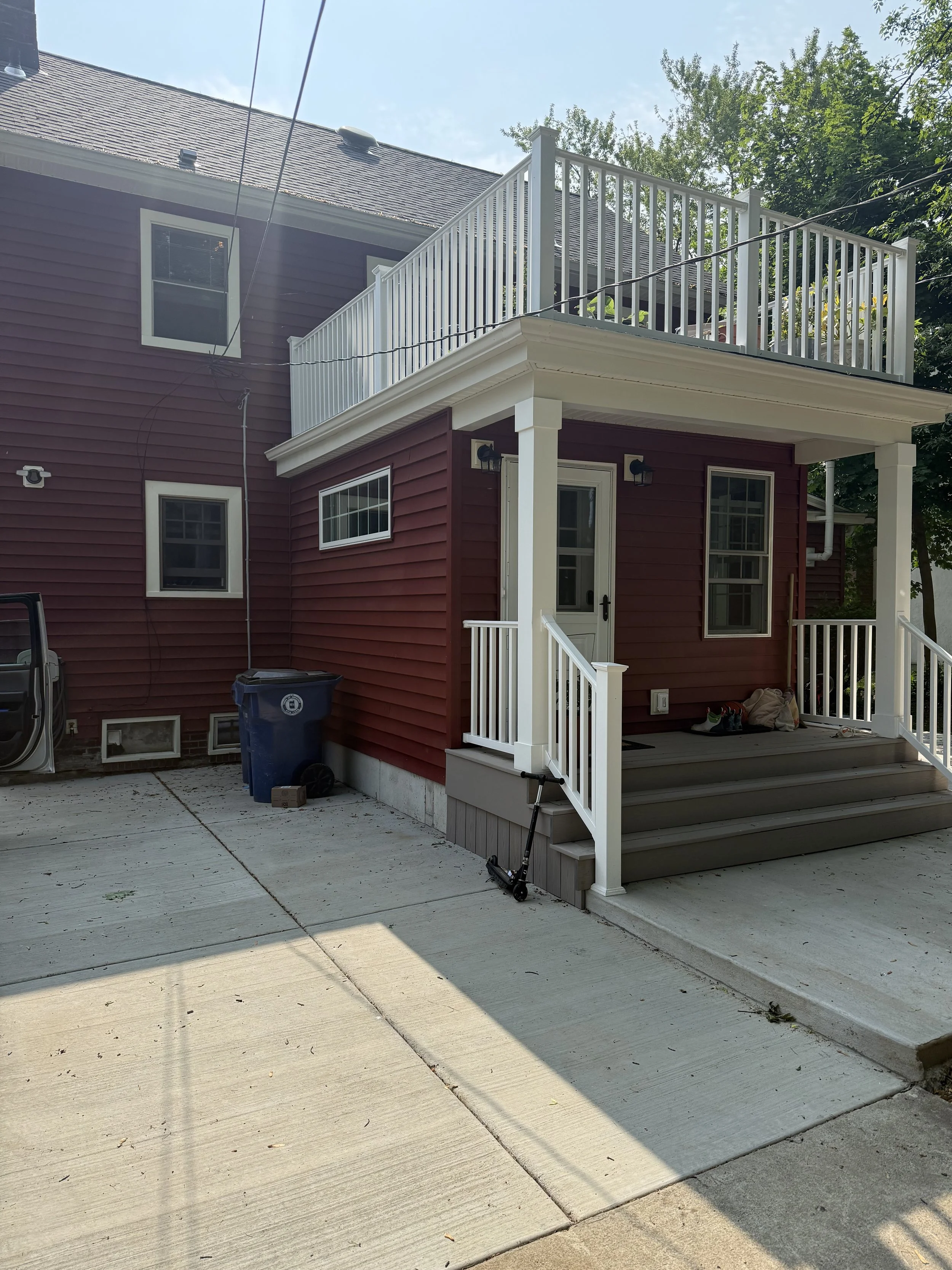 Back view of a two-story house with red siding and white trim, featuring a small porch with stairs and a balcony above. A scooter is leaning against the porch railing, and a few pairs of shoes are on the porch floor. A trash can is next to the house,