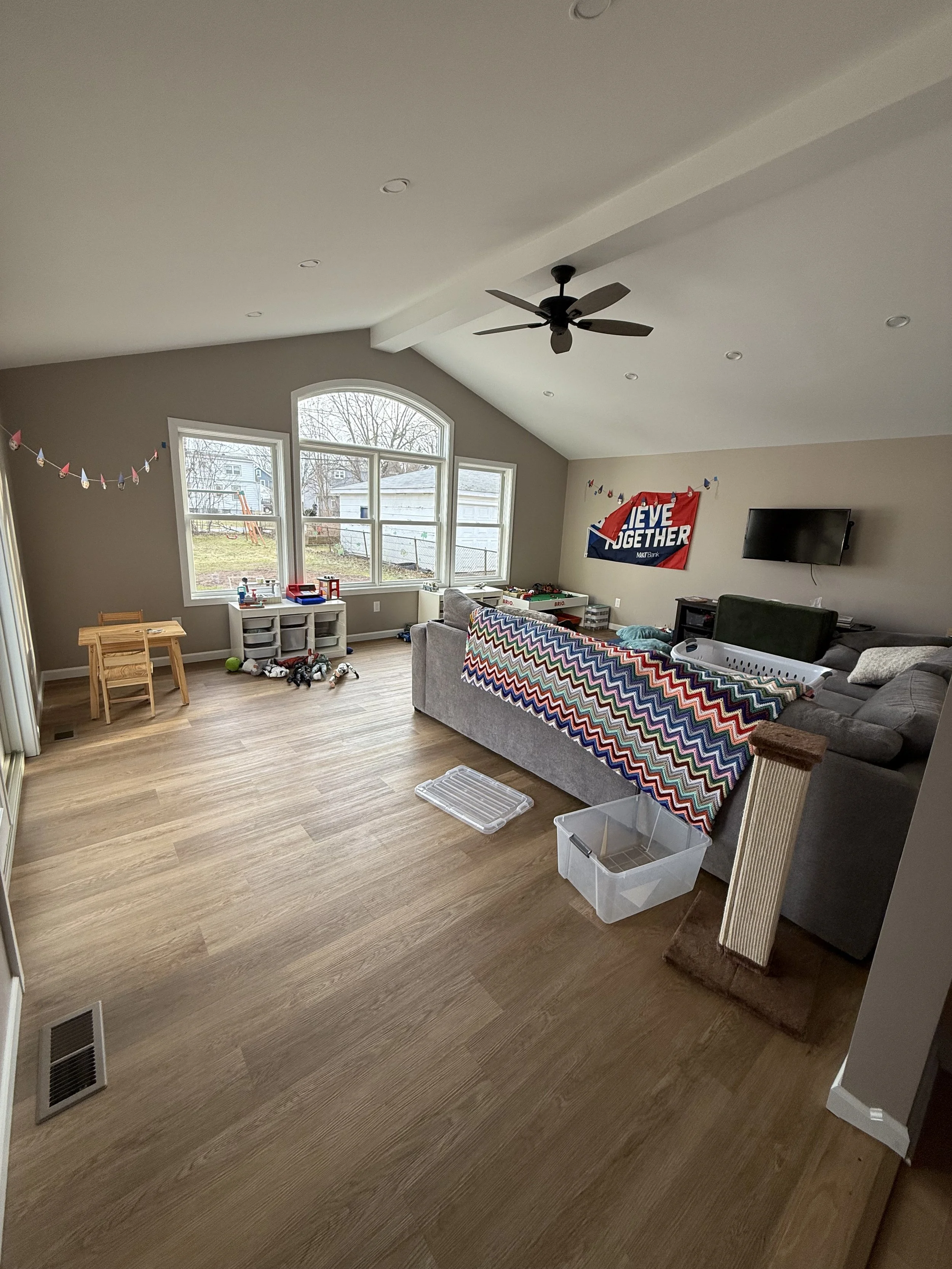 Living room with large windows, wooden floor, gray sectional sofa with a multicolored blanket, wall decorations including a Politically-themed banner, a TV, various toys and items scattered around.