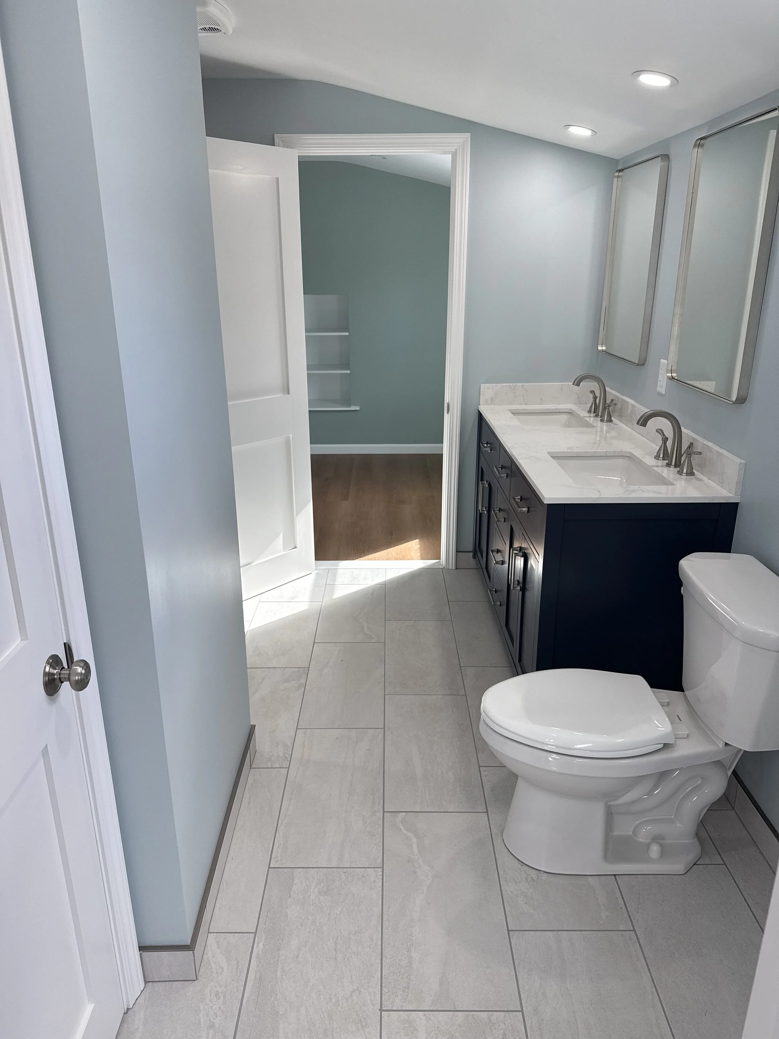 Bathroom with a double vanity with two mirrors, a toilet, and an open door leading to another room with wood flooring.