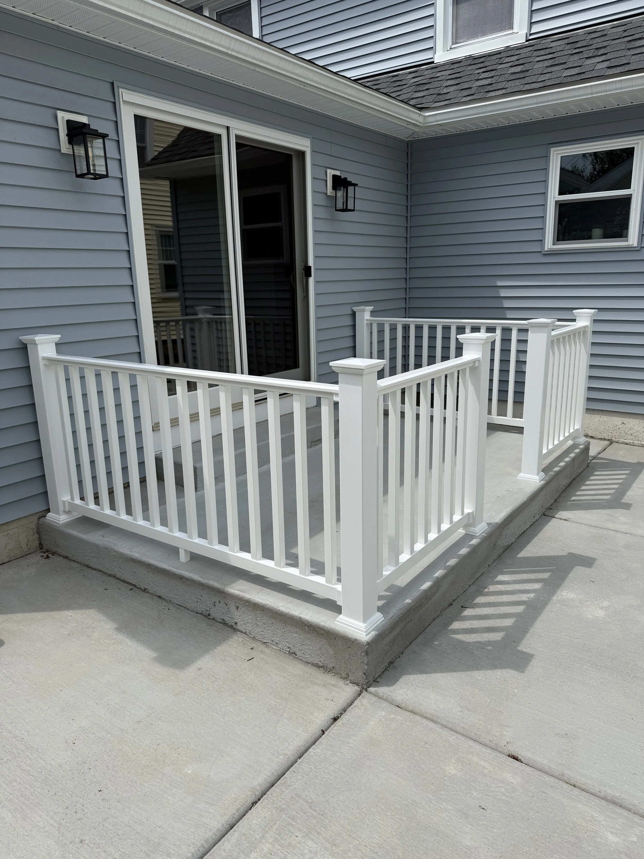 Backyard patio with a small white railing on a concrete surface, attached to a house with gray siding and black outdoor wall lanterns.