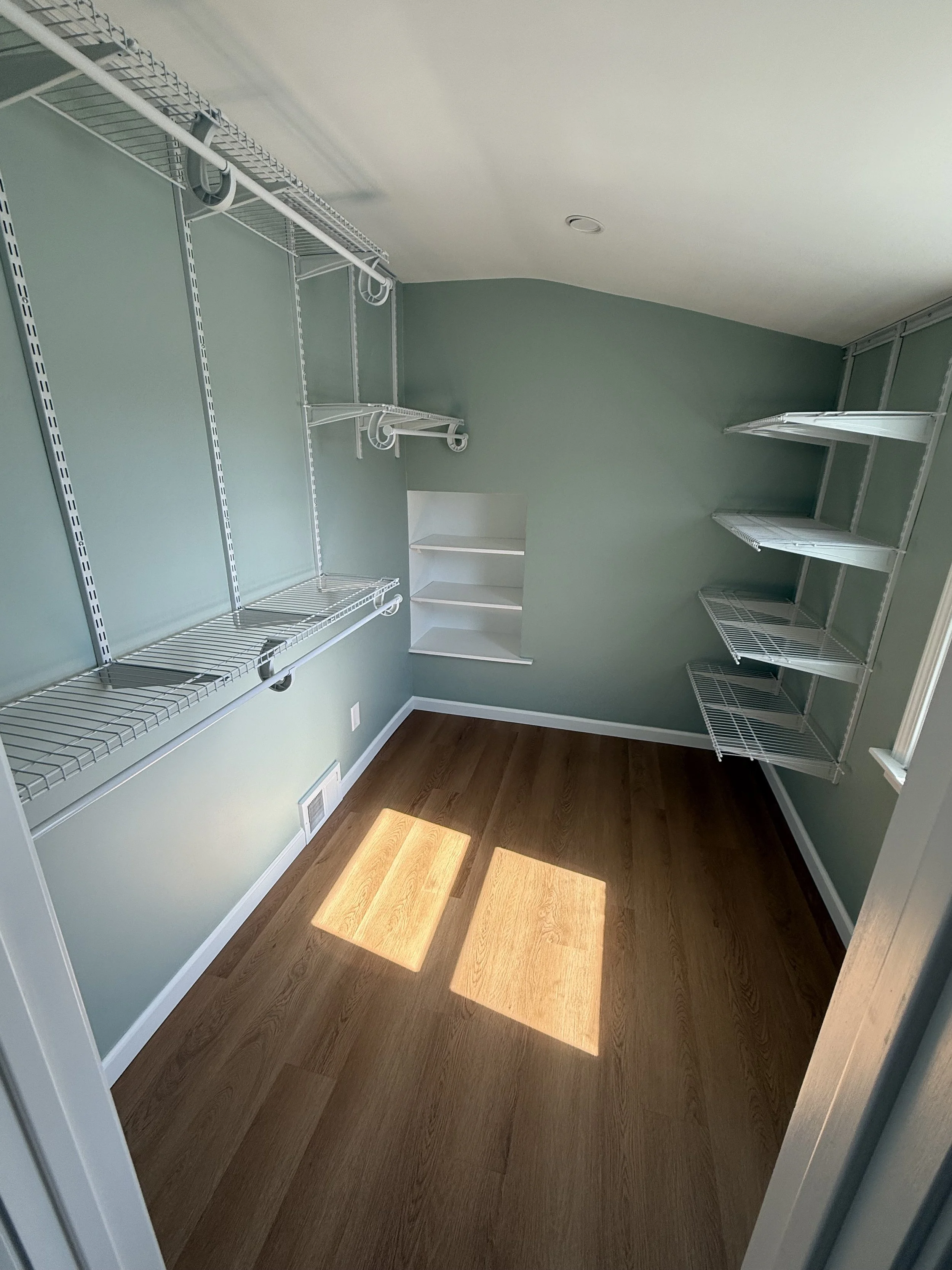 Empty walk-in closet with green walls, white wire shelving on three walls, and sunlight creating two patches of light on the wooden floor.
