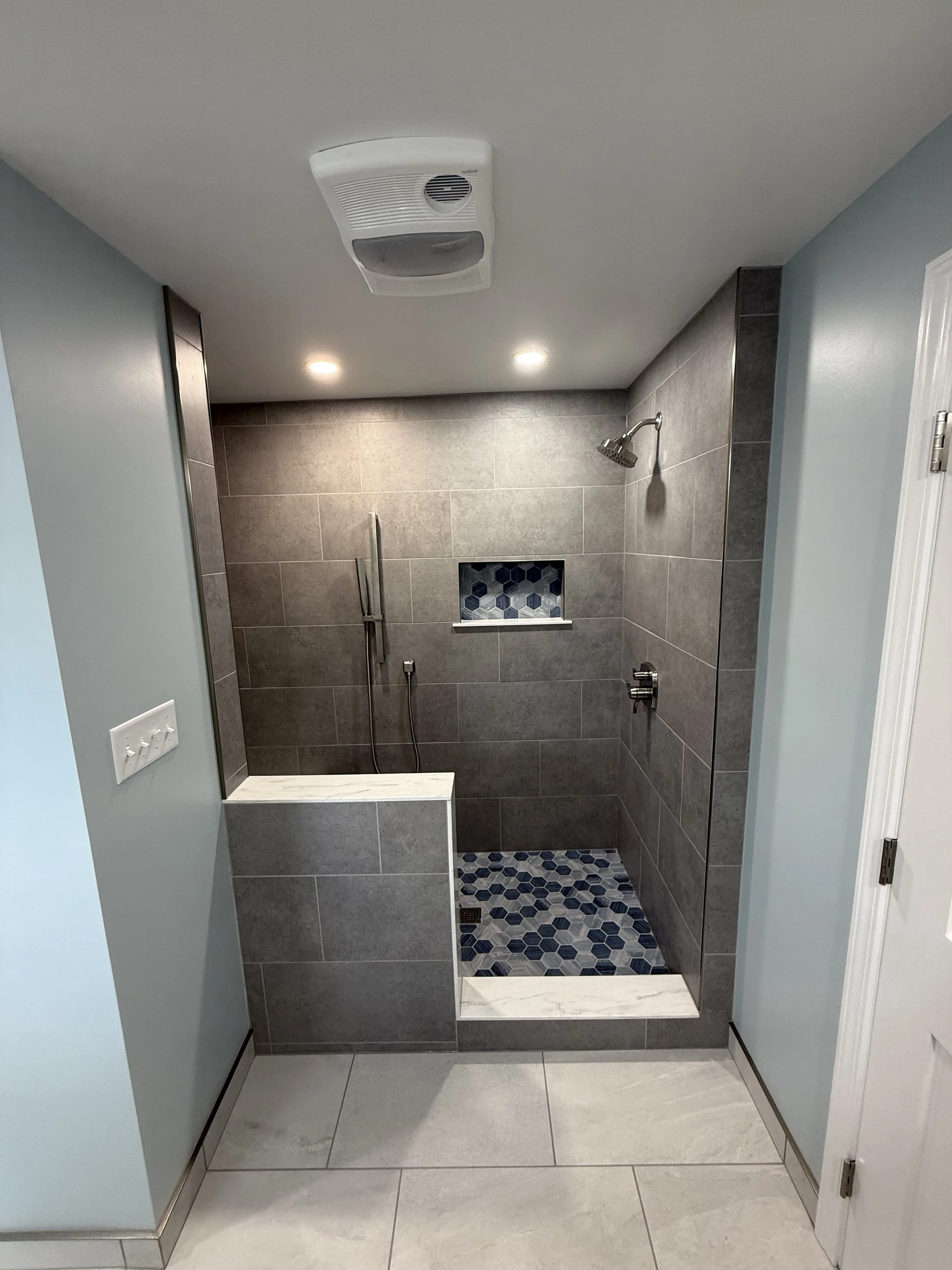 Bathroom shower with gray tiles, blue and white hexagonal floor tiles, small shelf with hex pattern, and chrome fixtures.