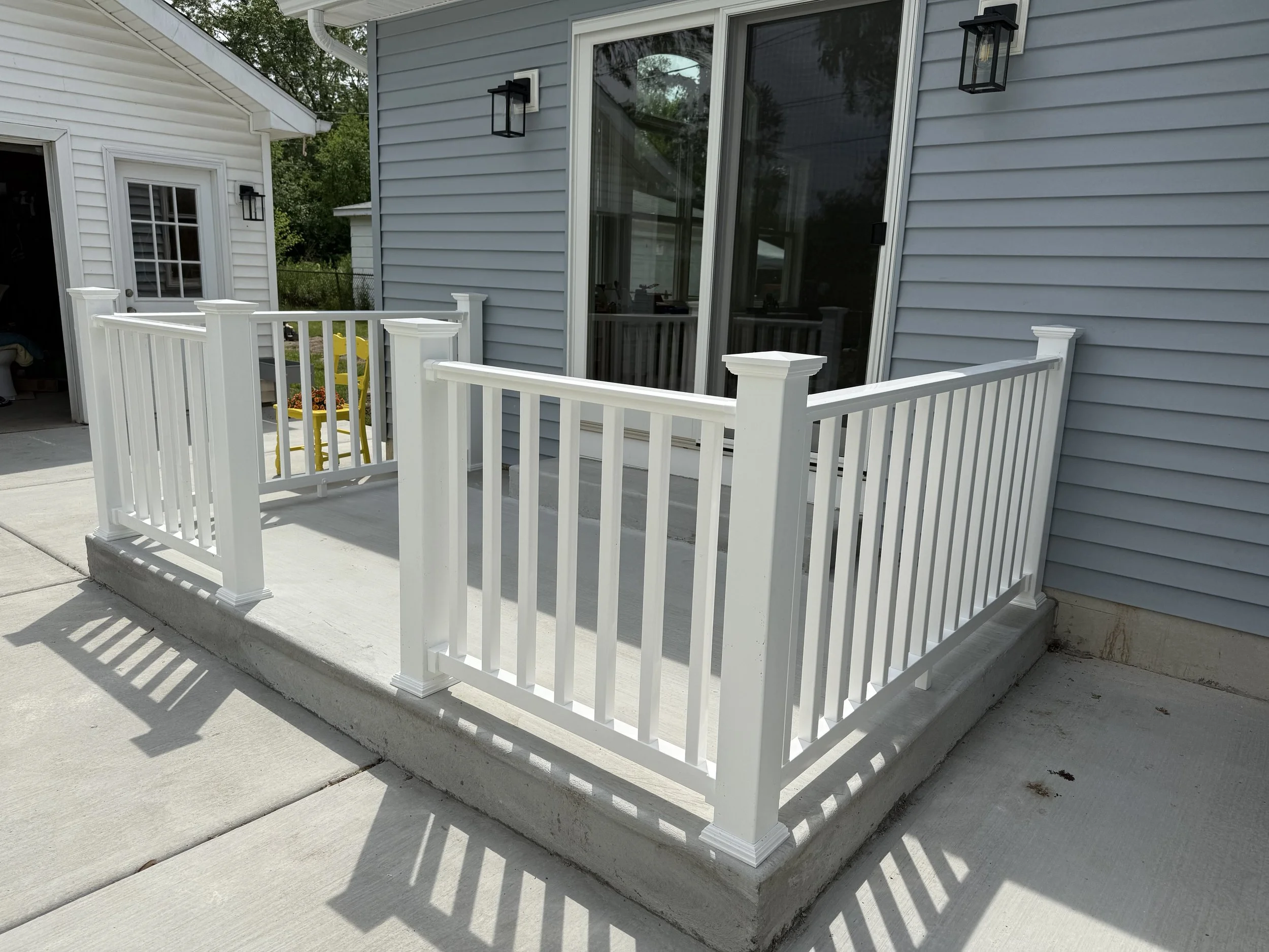 A small white deck with a white railing surrounding it, attached to the side of a house with gray siding. The deck has a concrete foundation and is located on a concrete patio.