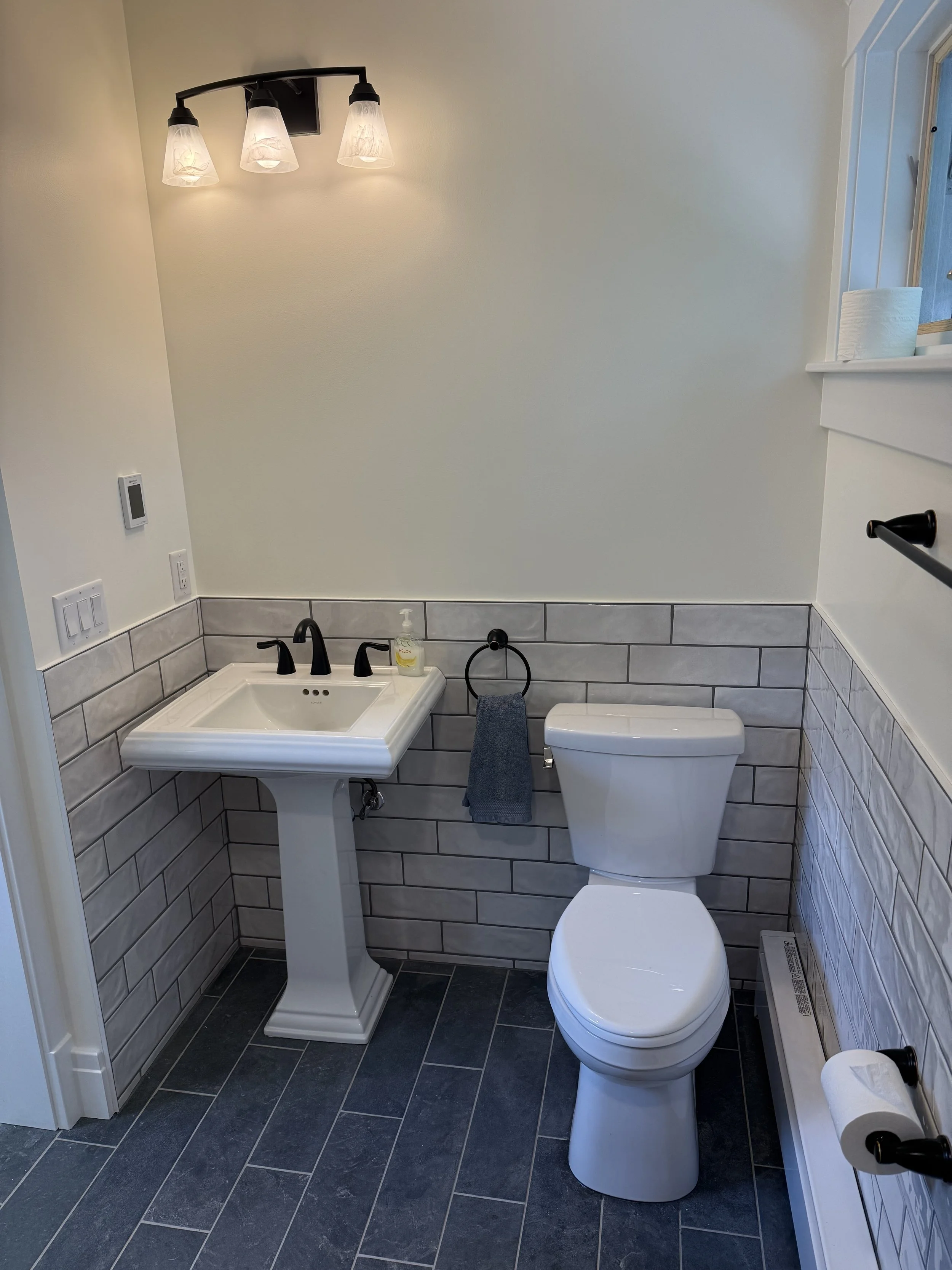 A small bathroom with a white toilet, a pedestal sink with black fixtures, a soap dispenser, a round mirror, black towel ring with a gray towel, black towel bar, tiled walls and floor, and a modern light fixture with three downward-facing glass shade