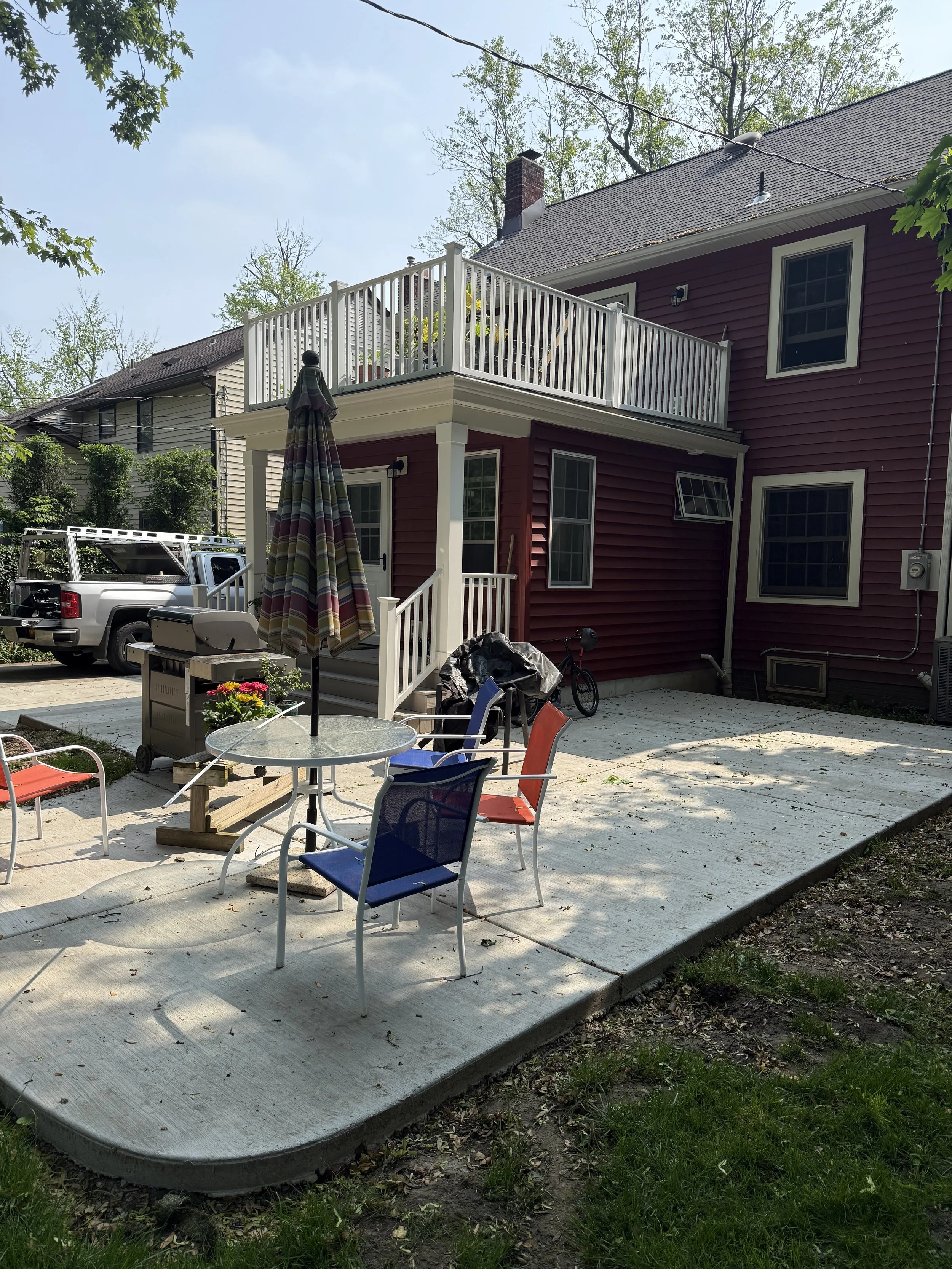 Backyard patio with outdoor furniture, a grill, and a bicycle, with a red house featuring a second-story balcony in the background.
