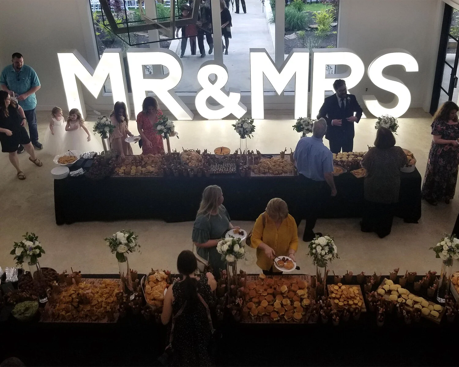 The main area of Lynn's Landing Venue indoor area is filled with two beautifully decorated buffet tables with family and friends gathered around. There is a large "Mr & Mrs" sign from Blossoms and Events in the background.