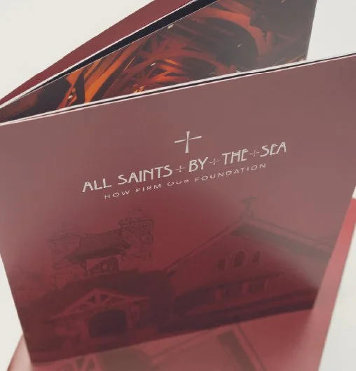 A pink book titled 'All Saints by the Sea: How FIRM Our Foundation' with a faint illustration of buildings on the cover.