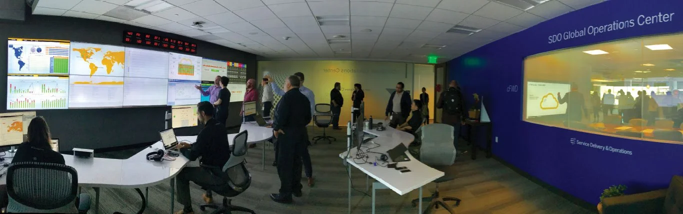 People working and monitoring in the SDO Global Operations Center with multiple screens displaying data and maps, and a window showing additional staff.