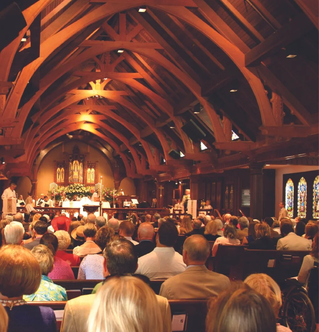 People gathered in a church with wooden vaulted ceilings for a service or ceremony, with an altar decorated with flowers at the front and stained glass windows on the sides.