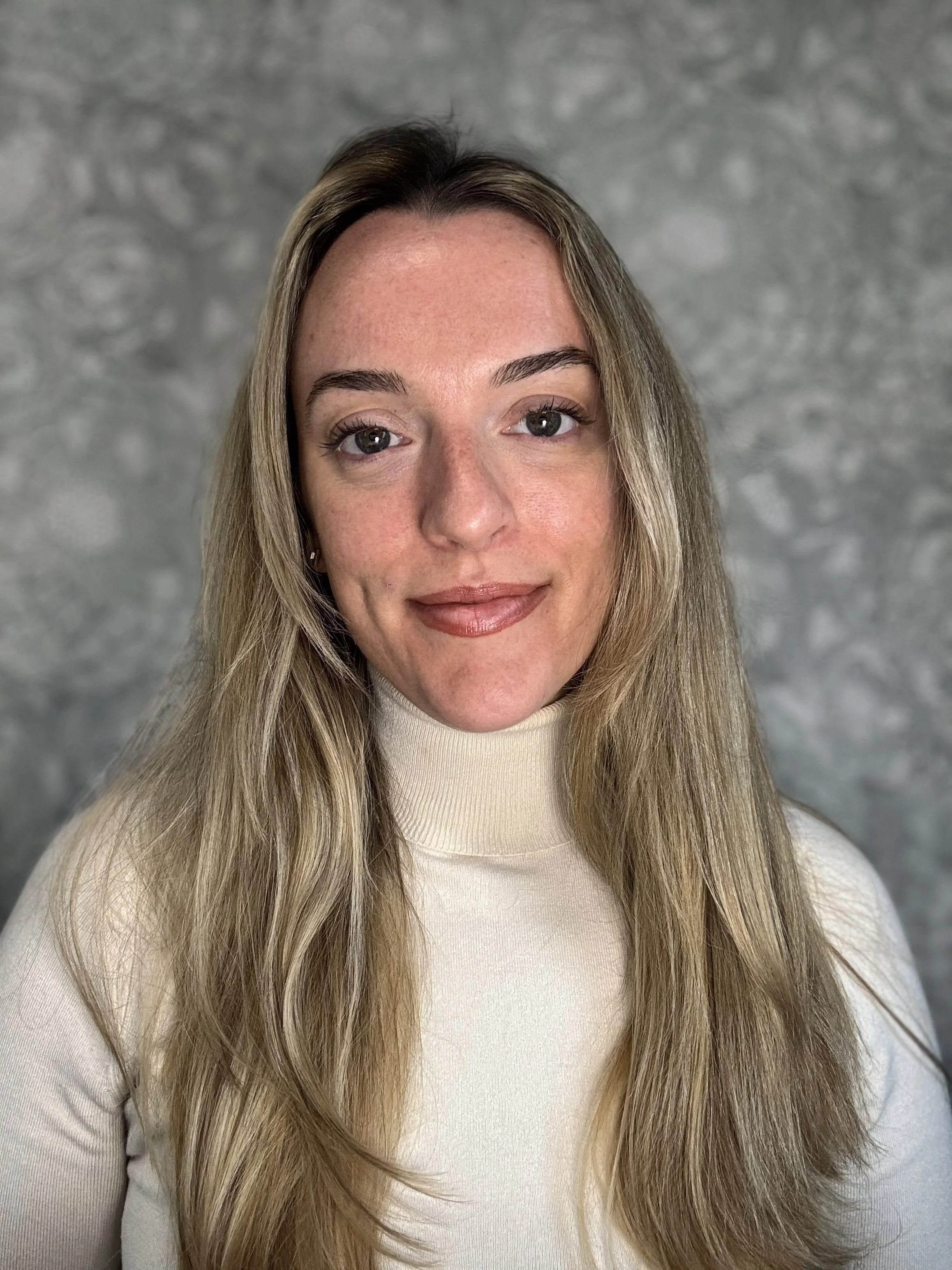 Portrait of a woman with long blonde hair, wearing a cream turtleneck sweater, standing in front of a gray textured background.