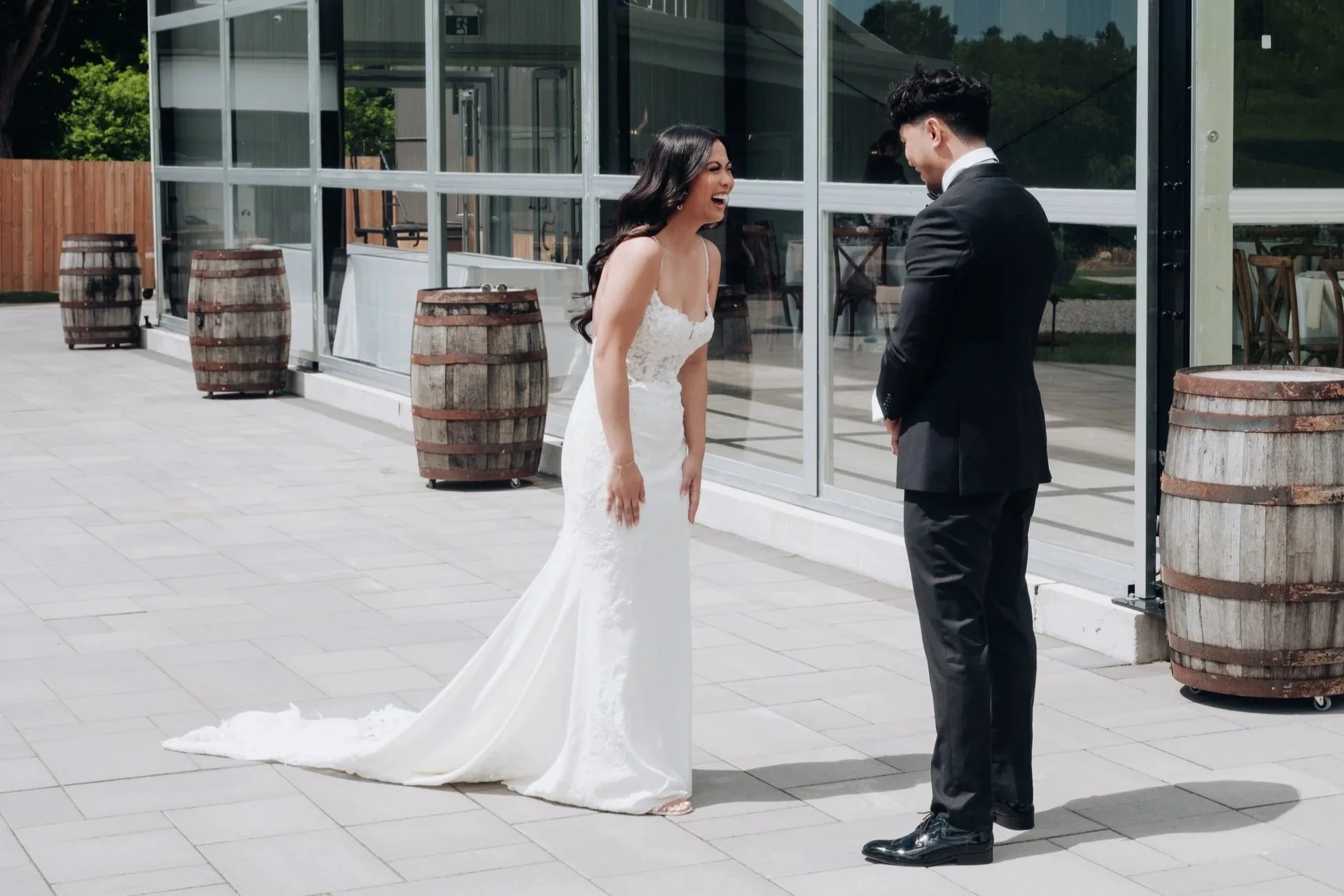 Bride and groom sharing a happy moment outside a glass-walled building during their wedding, with the bride in a white lace gown and the groom in a black suit.