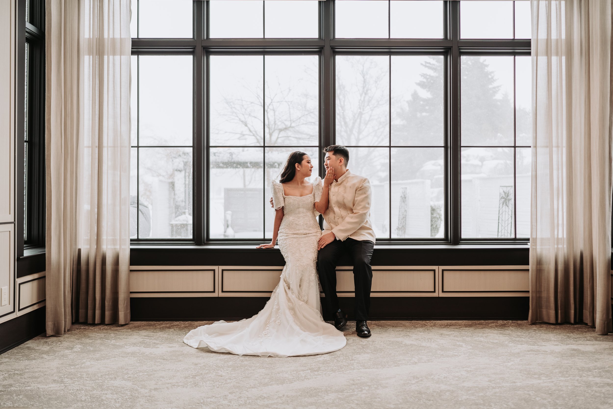 A bride and groom sitting on a window seat during winter, gazing at each other. The bride is wearing a white lace gown, and the groom is in a cream-colored suit jacket and black pants. Snow can be seen outside the large window behind them.