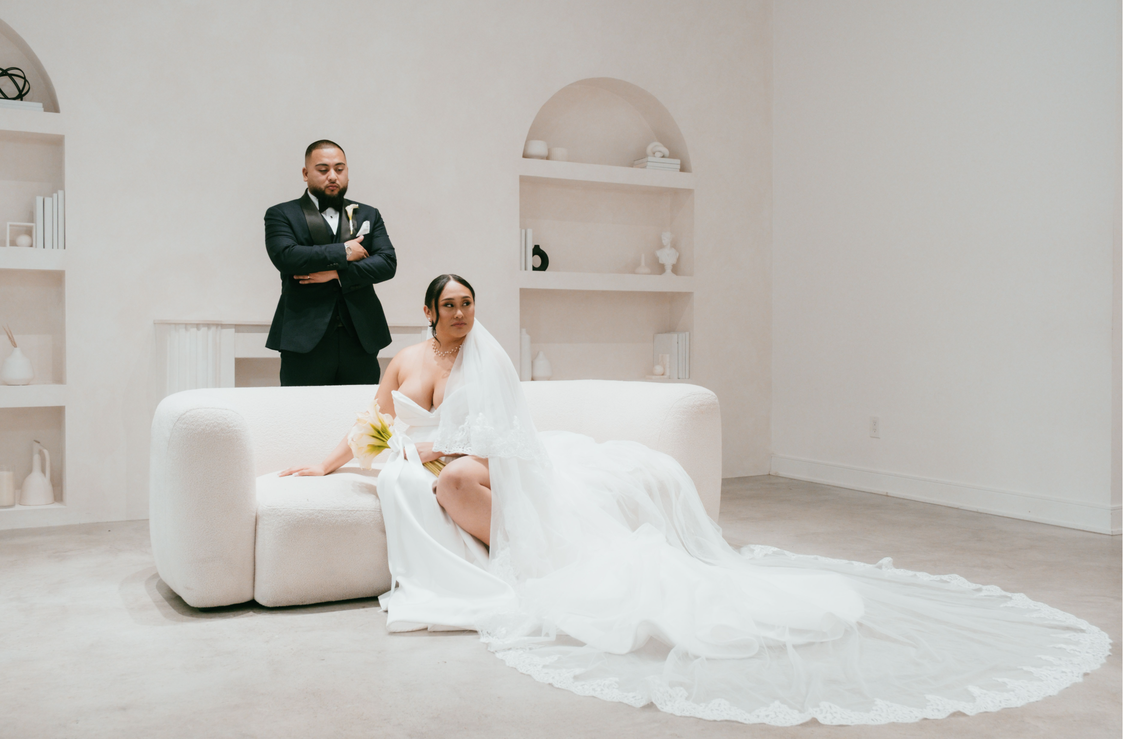 Bride in a white wedding dress sitting on a white couch, groom in a tuxedo standing behind her with arms crossed, minimalist white room with built-in shelves and neutral decor.