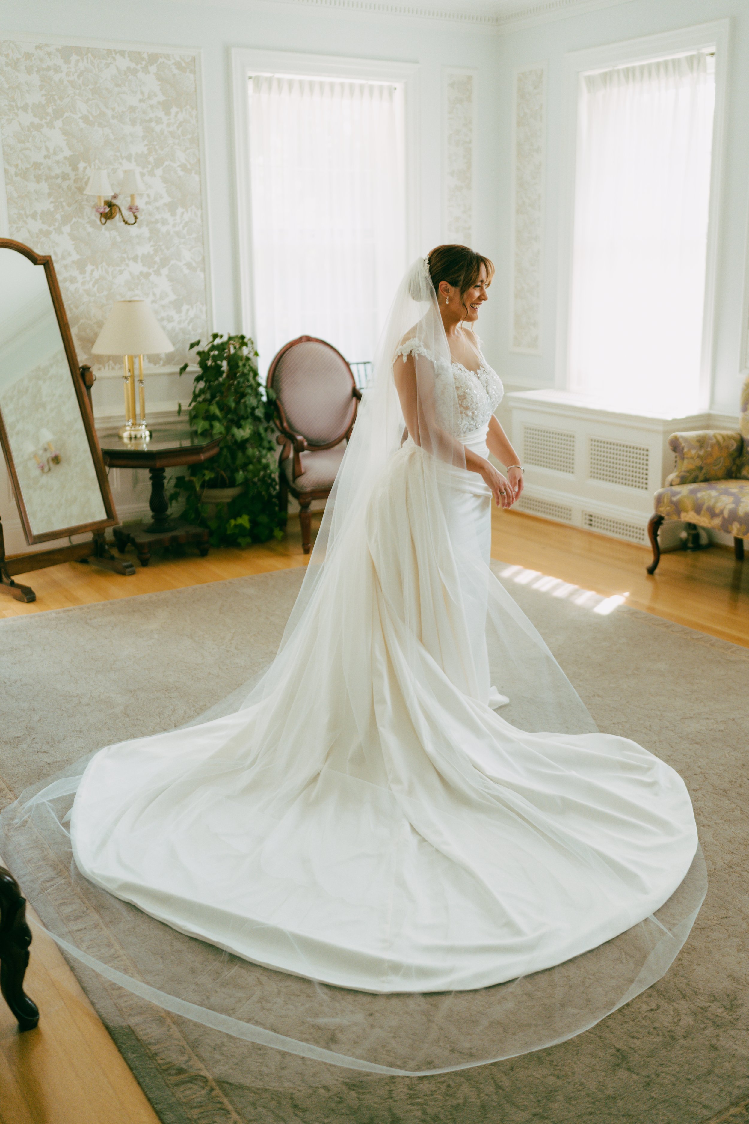 A bride in a wedding dress standing in a well-lit room with large windows, a mirror, a chair, and a lamp.