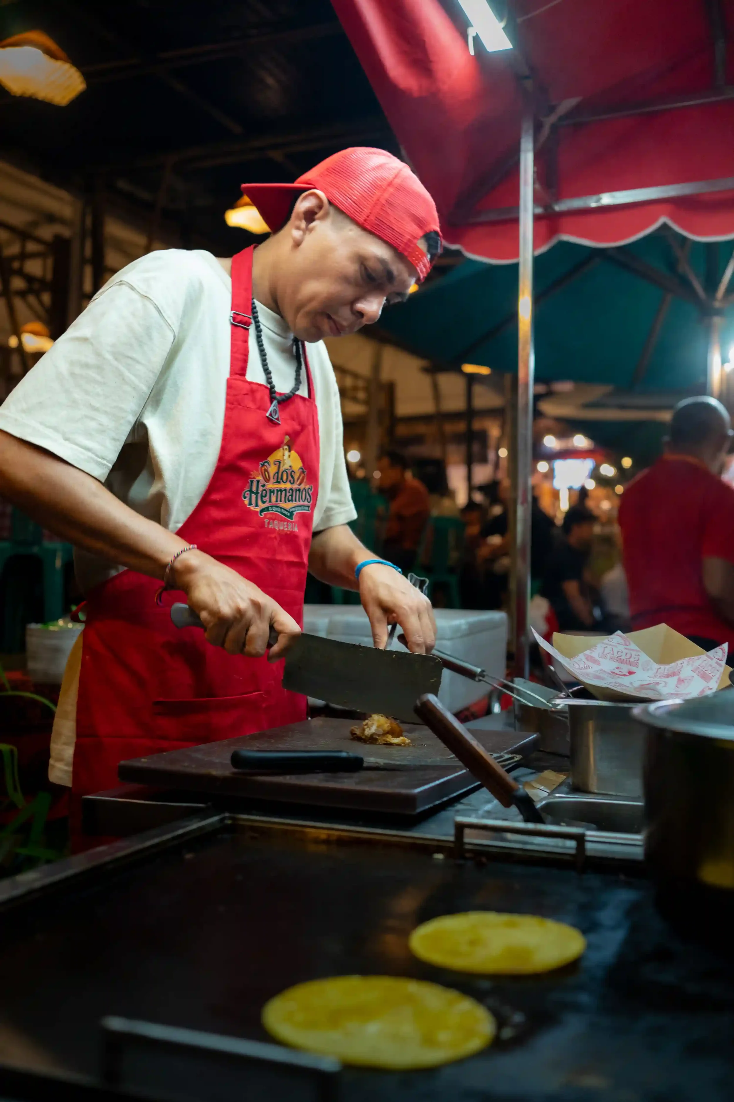A street food vendor wearing a red apron chops meat with a large cleaver at a busy taco stand in Mexico, preparing ingredients on a wooden cutting board.